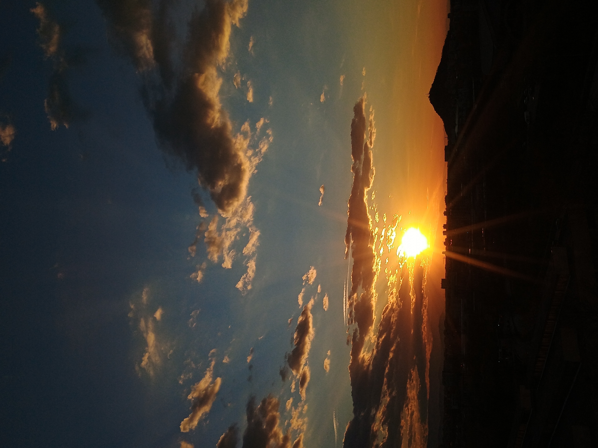 The image depicts a sunset in Plovdiv, Bulgaria, with the sun setting over buildings. The sky is a gradient of blue to orange, with clouds scattered throughout. The sun is partially obscured by clouds, but still shines brightly. There are rays of light emanating from the sun. The buildings are silhouetted against the bright sky. The overall scene is peaceful and serene.