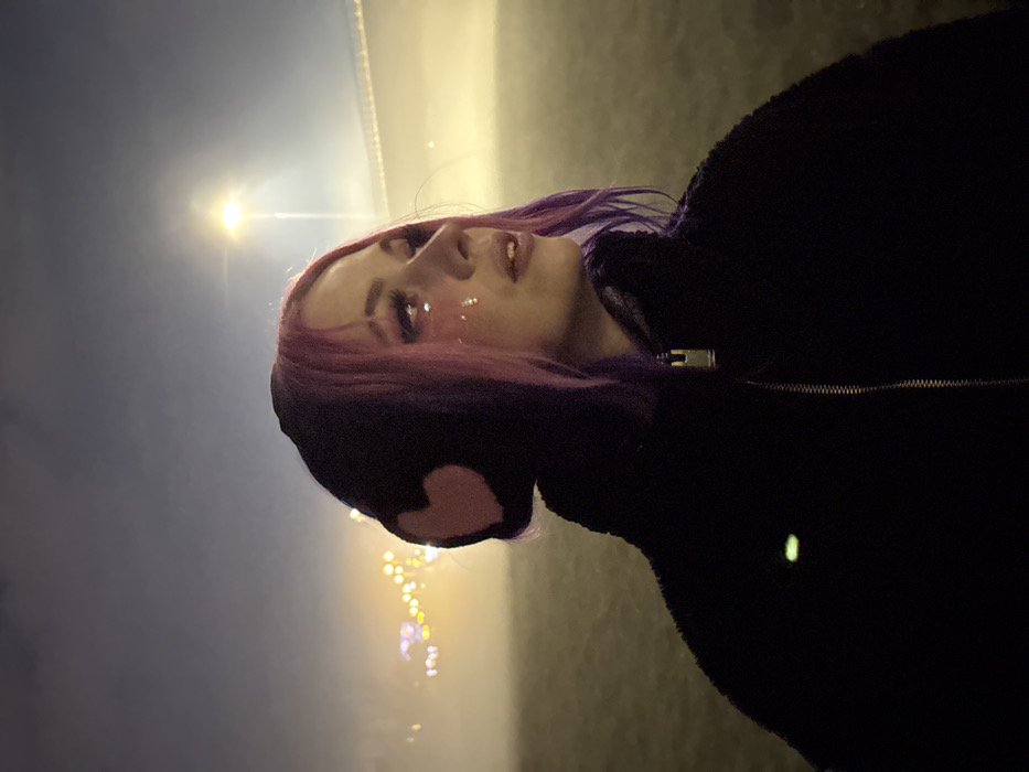 A young adult female is depicted outdoors at night, likely situated along the banks of the Sava River in Zagreb, Croatia. The subject is positioned off-center to the left, shown in profile with her head turned to the right, and her gaze directed slightly upwards. She has shoulder-length hair, dyed in purple and pink shades, and a cluster of iridescent glitter applied to her right cheekbone below the eye. She wears a black, textured, zip-up jacket with a silver-colored zipper pull and a black beanie hat adorned with a prominent pink heart emblem covering her right ear. The environment is characterized by heavy fog or haze, diffusing all light sources. In the background, beyond a wide, dark expanse of water, the illuminated superstructure of the Most Slobode (Liberty Bridge) is visible, accompanied by distant, blurred city lights and a solitary bright streetlamp. The immediate foreground consists of a dark, grassy area.