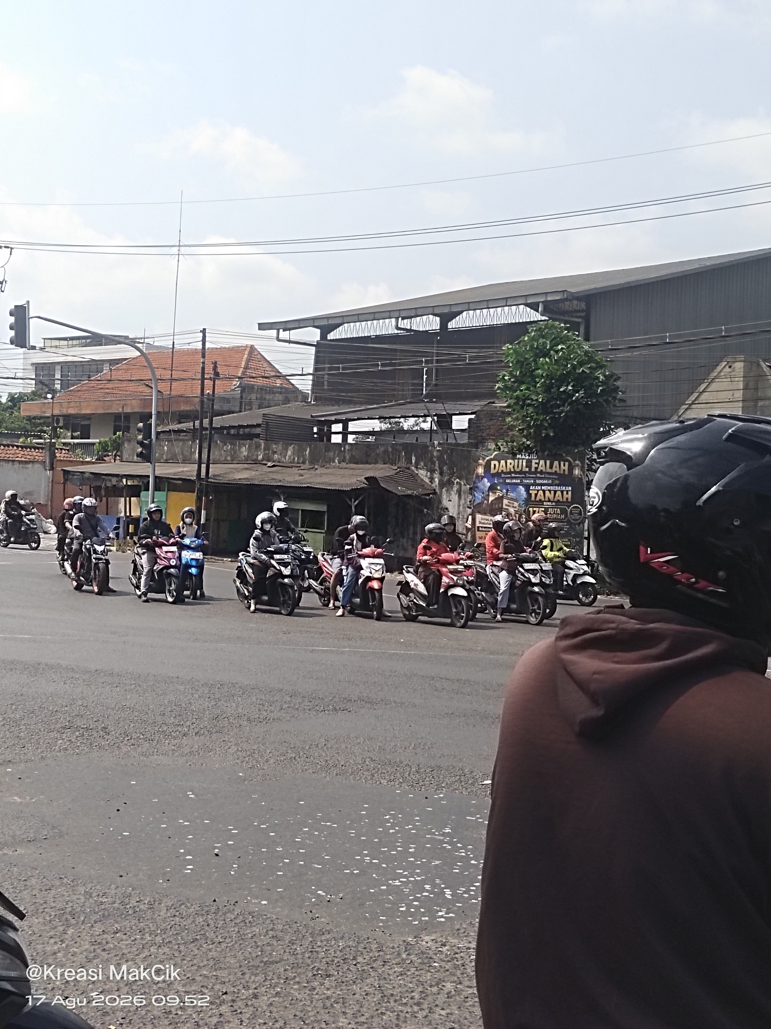 A group of motorcyclists, all wearing helmets and most wearing masks, are proceeding through an intersection in what appears to be a commercial or industrial area. The motorcyclists are on a paved road, and traffic signals are visible. In the background, there are buildings with corrugated metal roofs, as well as a large billboard. The sky is partly cloudy. The timestamp at the bottom of the image reads "17 Agu 2026 09.52" and a watermark says "@Kreasi MakCik".