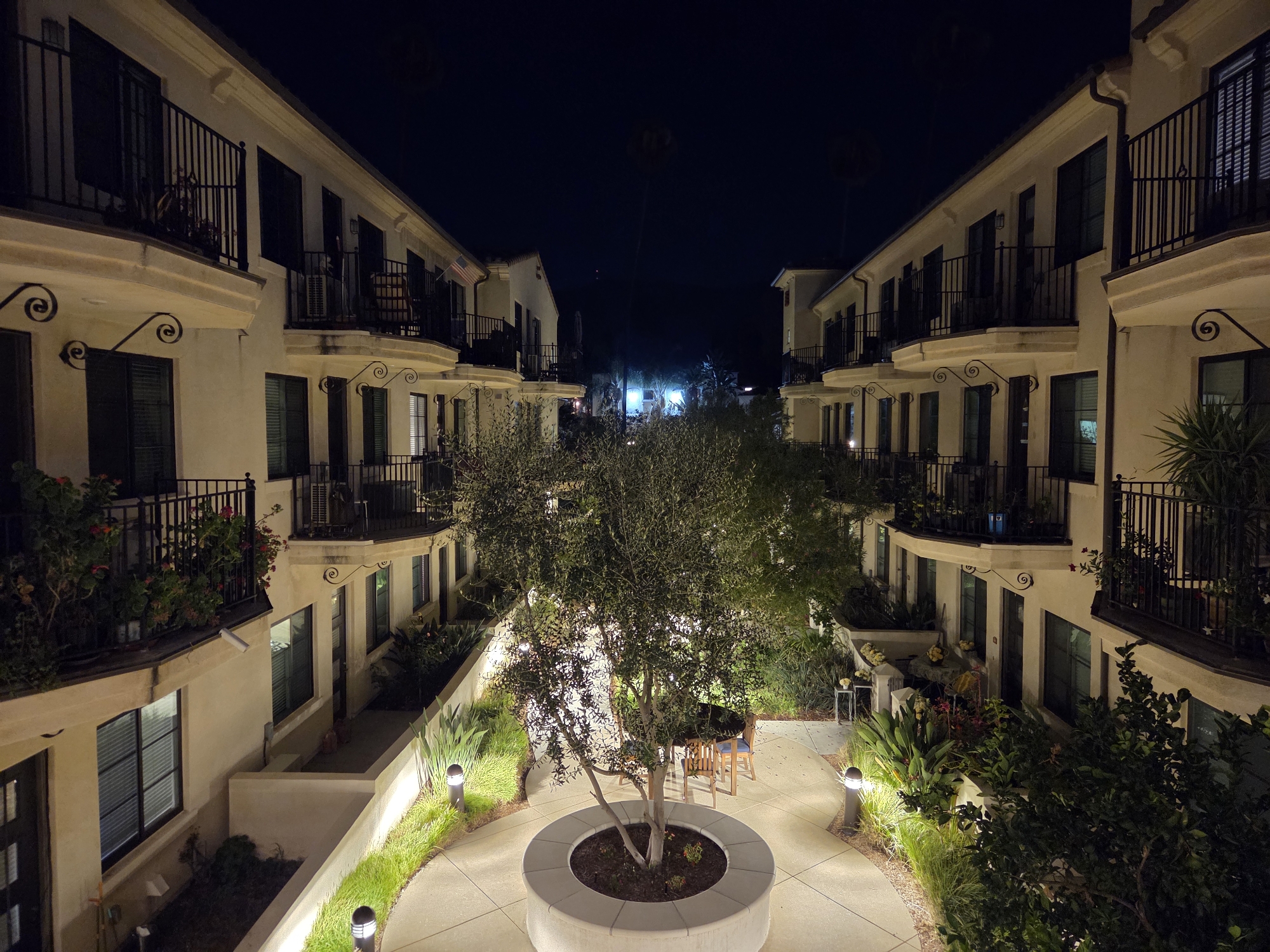 The image captures a view of a courtyard at night in Glendale, California, likely part of a residential complex. Two multi-story buildings with cream-colored stucco exteriors and Spanish-style tile roofs face each other across a paved courtyard. Both buildings feature balconies with black wrought-iron railings on the upper floors, adorned with potted plants and some outdoor furniture visible on one balcony. Windows with dark shutters are interspersed along the building facades.

In the center of the courtyard is a circular raised planter containing a small, mature tree with visible branches and green foliage. Around the tree, a circular concrete seating area is present, and beyond that, a dining table with four chairs is set up. The courtyard floor is paved with large concrete tiles. Low-lying green plants and ornamental grasses line the edges of the courtyard pathways, illuminated by modern, cylindrical bollard lights. The background reveals a dark night sky with some distant lights and palm trees. The overall scene conveys a sense of quiet evening ambiance.
