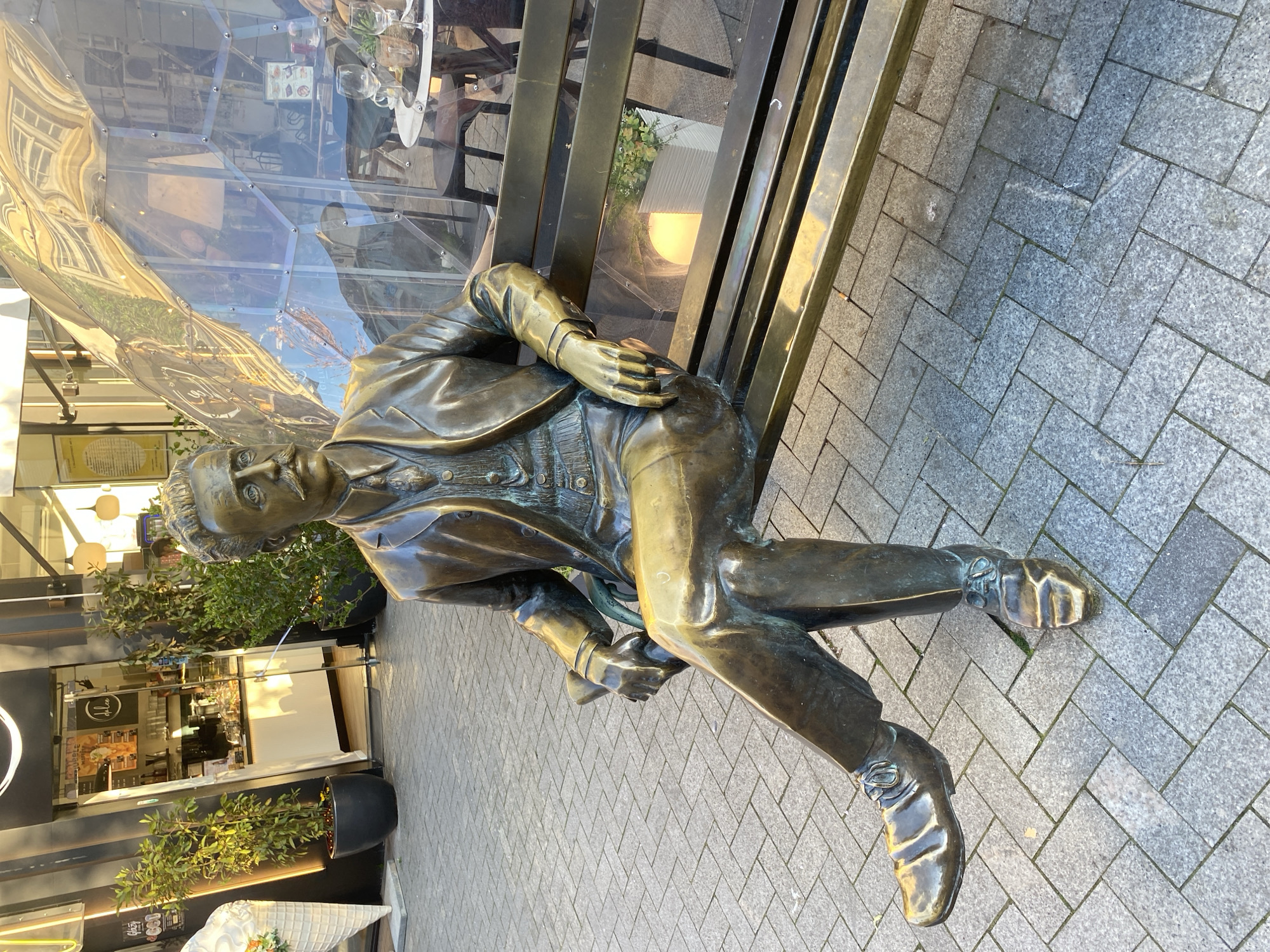 A bronze sculpture of a seated man on a bench is located outside a café or restaurant. The statue appears to be a historical figure, dressed in a suit. The sculpture is seated on a bronze bench set against a building with reflective glass panels. The café's interior is visible through the glass. The ground is paved with grey herringbone pattern tiles. The statue is located in Varna, Bulgaria.