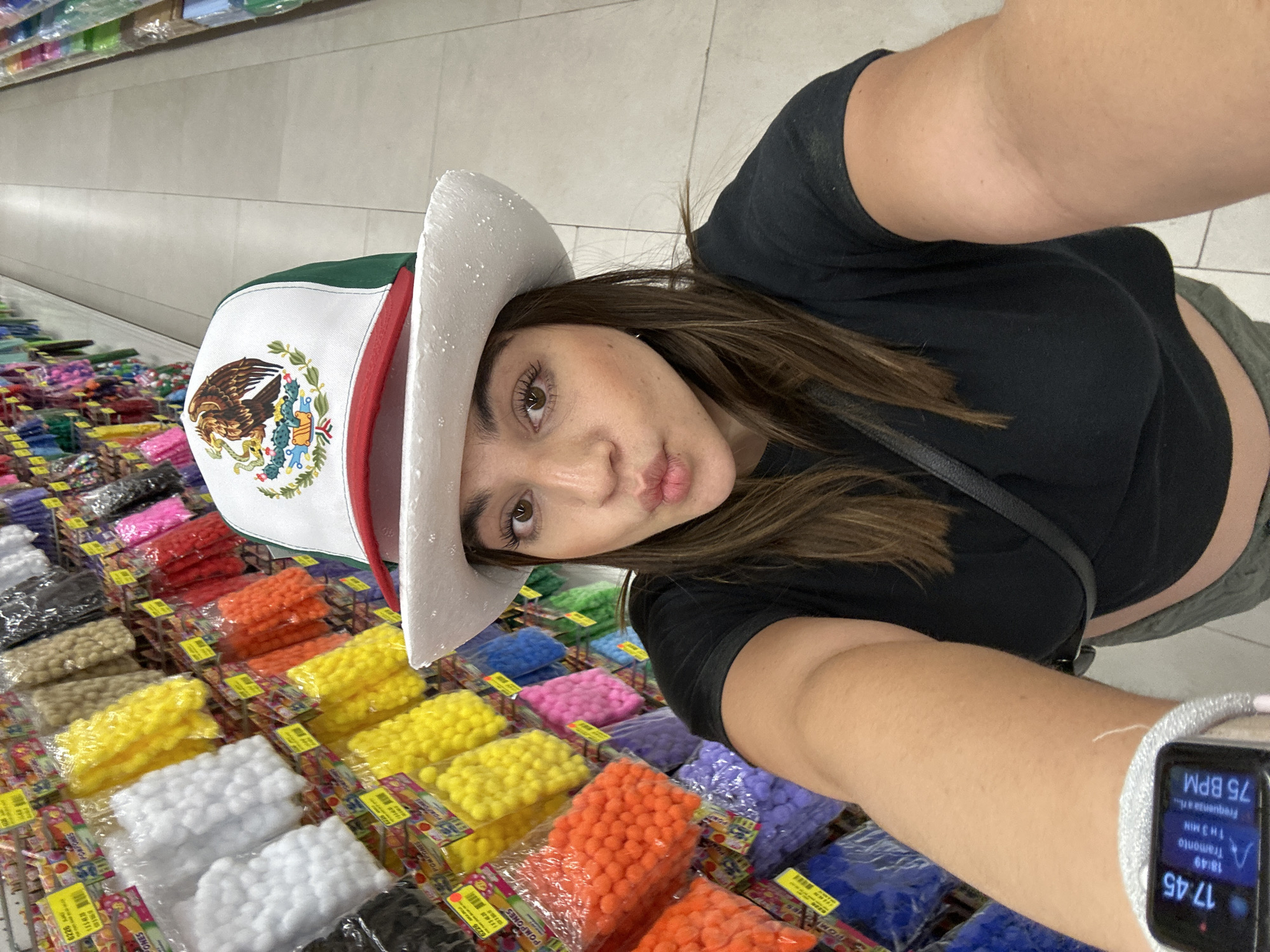The image shows a woman taking a selfie. She is wearing a black shirt and a white cowboy hat with a Mexican flag emblem on the front. Her lips are puckered. A white watch is visible on her wrist. In the background is a colorful display of pom-poms in plastic packaging. Based on the flag emblem and the OCR of "POMPONES" on the product packaging, the location is likely in Mexico. Given the information in the OCR, it is most likely that the city name is "San Bernardino Tlaxcalancingo, Mexico".