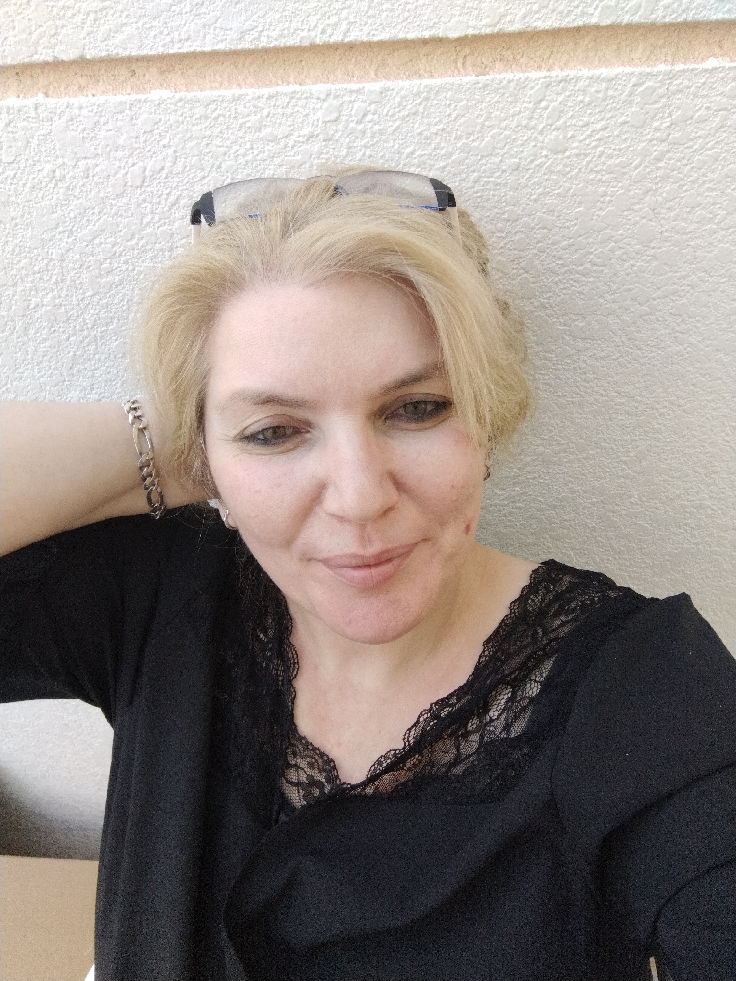 A medium shot captures a woman with blonde hair, wearing a black top with lace detailing around the neckline. She has on a silver bracelet on her left wrist and her left arm is bent, with her hand resting behind her neck. She is looking towards the camera with her head slightly tilted. She is wearing glasses pushed up on her head. The background is a textured, light-colored wall with a horizontal stripe. The scene appears to be outdoors, as suggested by the lighting and the textured wall, which is common in exterior architectural elements in regions like Al Mas'oudi, United Arab Emirates.
