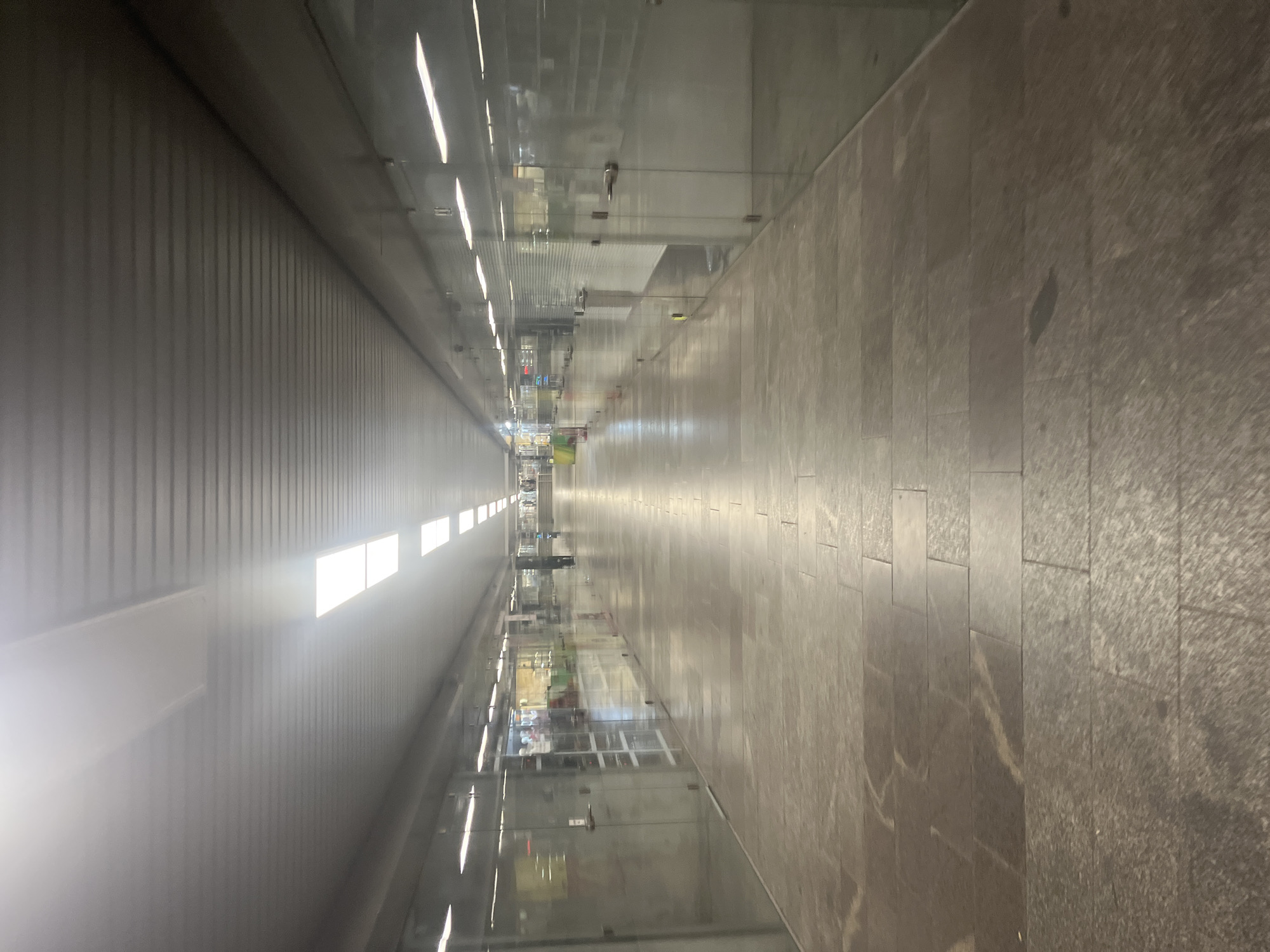 The image depicts a long, brightly lit corridor in Plovdiv, Bulgaria. The corridor appears to be part of an underground passage or pedestrian walkway. The left wall is constructed of metallic gray panels with horizontal rows of rectangular lights. The right side features glass walls, likely storefronts, reflecting the corridor's lights and ceiling. The floor is composed of dark gray stone tiles, creating a textured, slightly worn surface. A few distant figures are visible towards the end of the corridor. The perspective emphasizes the length and linearity of the space.