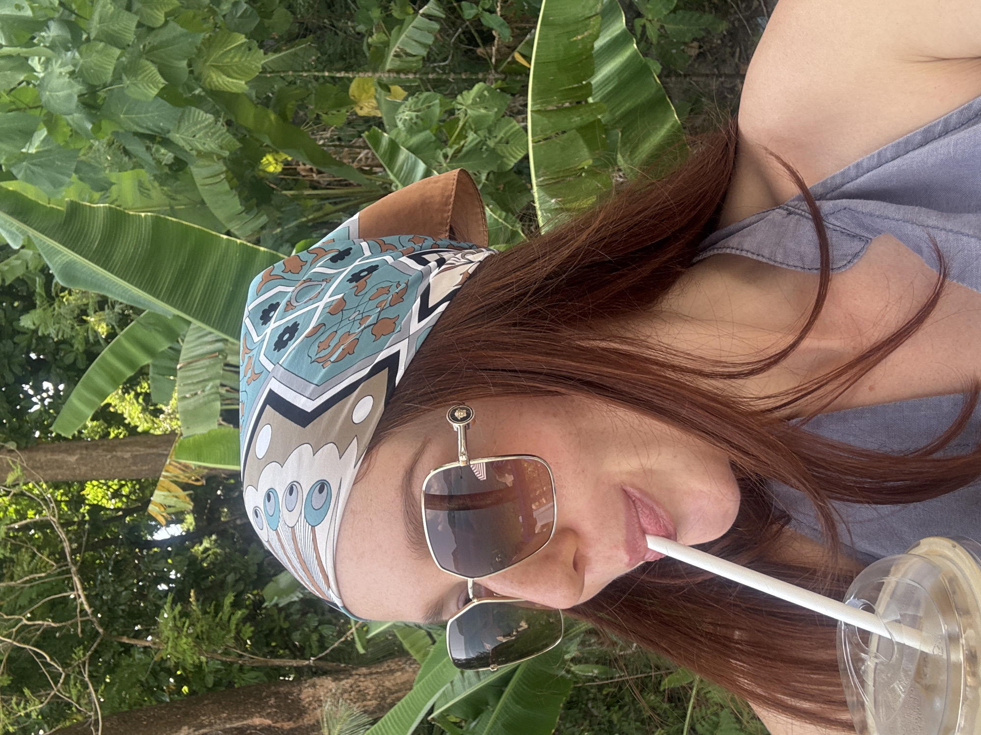 A red-haired woman wearing a patterned headscarf and sunglasses is seen sipping from a plastic cup with a straw. She is wearing a gray top. The background shows green tropical vegetation, specifically banana leaves. The location is Kamala, Thailand.
