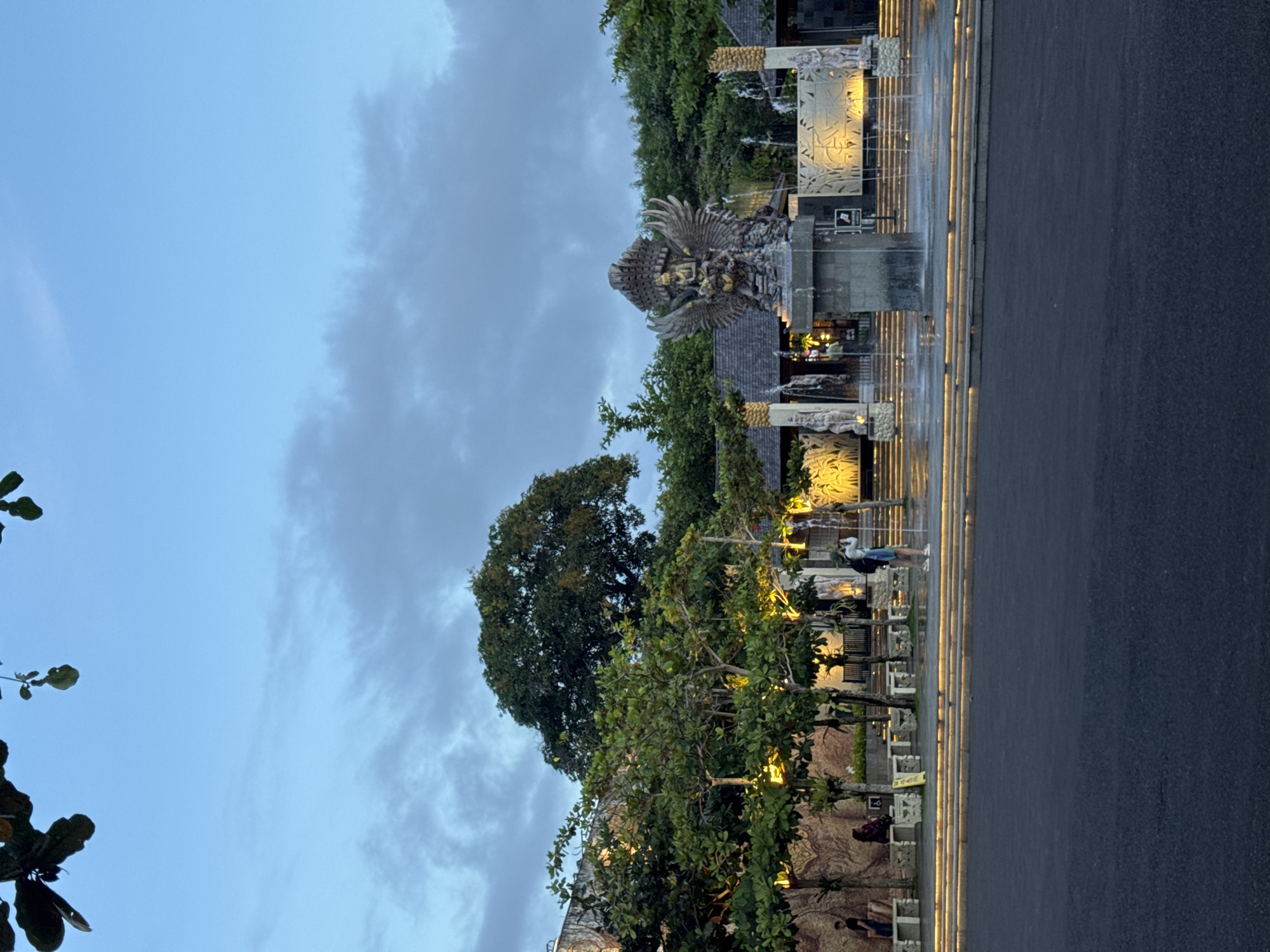 This image captures an evening scene at a grand outdoor cultural park or plaza in Kuta Selatan, Indonesia, strongly resembling the Garuda Wisnu Kencana (GWK) Cultural Park.
The central subject is a towering, intricately detailed statue of a winged deity, likely Garuda, with outstretched wings, positioned prominently on the right side of the frame. This majestic sculpture is surrounded by several decorative fountains and water features, which reflect the soft glow of artificial lights.
Lush green trees and dense foliage are abundant throughout the scene, framing the structures and adding natural beauty. In the background, hints of traditional architectural elements and buildings can be seen beyond the trees and fountains.
A wide, dark paved area, possibly a road or plaza, spans horizontally across the lower portion of the image. This area is distinctly illuminated by warm, linear strip lights embedded along its edge, creating a visually appealing boundary. A few people are visible strolling or standing on the paved areas, enjoying the ambiance of the illuminated setting.
The time of day is dusk or early evening, evidenced by the gentle transition of the sky from a clear blue to softer gray tones with some visible clouds. The artificial lighting on the statue, fountains, and ground strips creates a warm and inviting atmosphere. The overall mood is peaceful and majestic. No legible text is visible within the image.