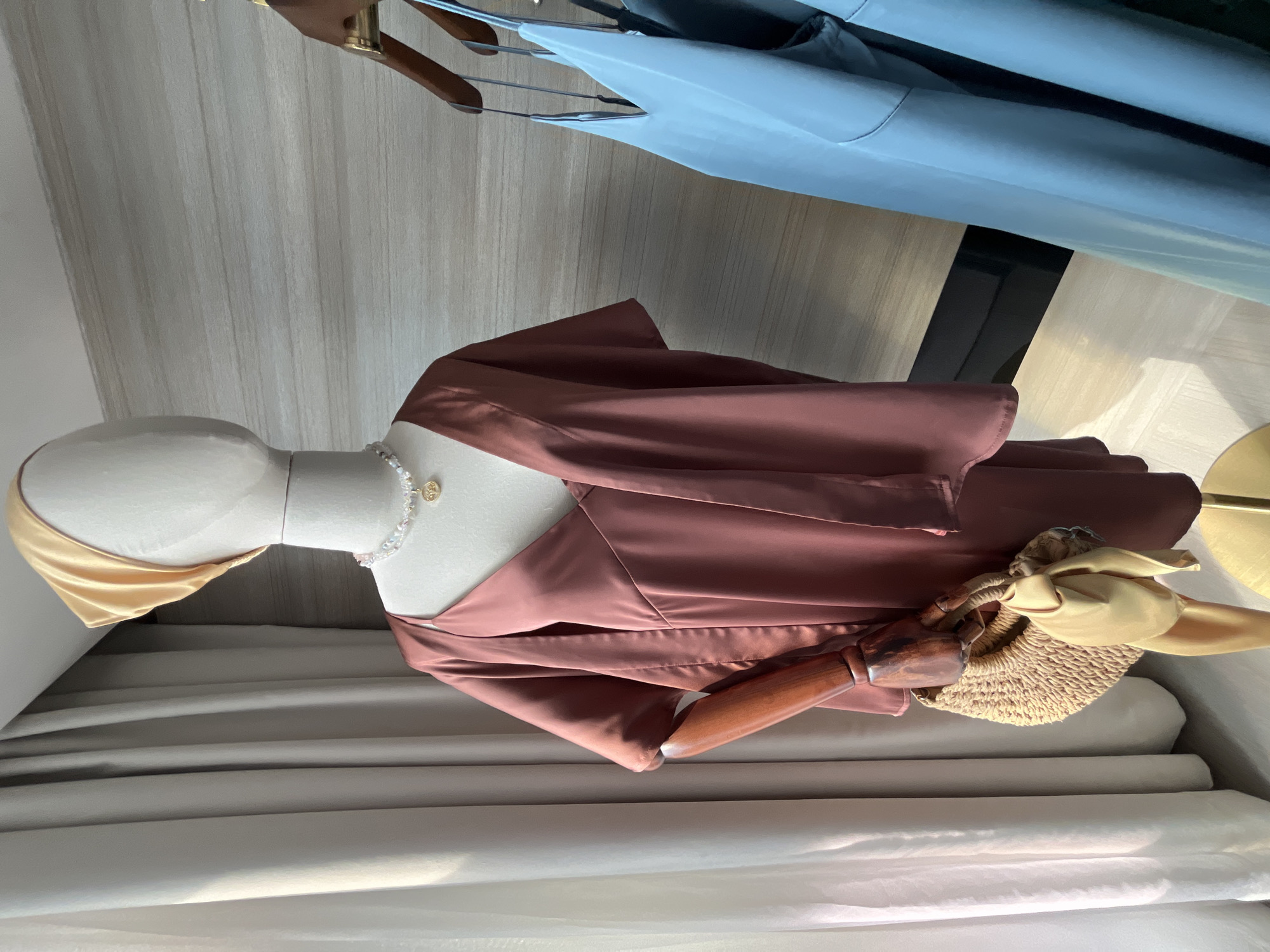 A mannequin stands in Bac Nha Trang Ward, Vietnam. It wears a brown silky outfit with a gold accessory and a pearl necklace. A woven bag hangs on its arm. Two dresses are hung on a rack in the background. A cream-colored backdrop is behind the mannequin.