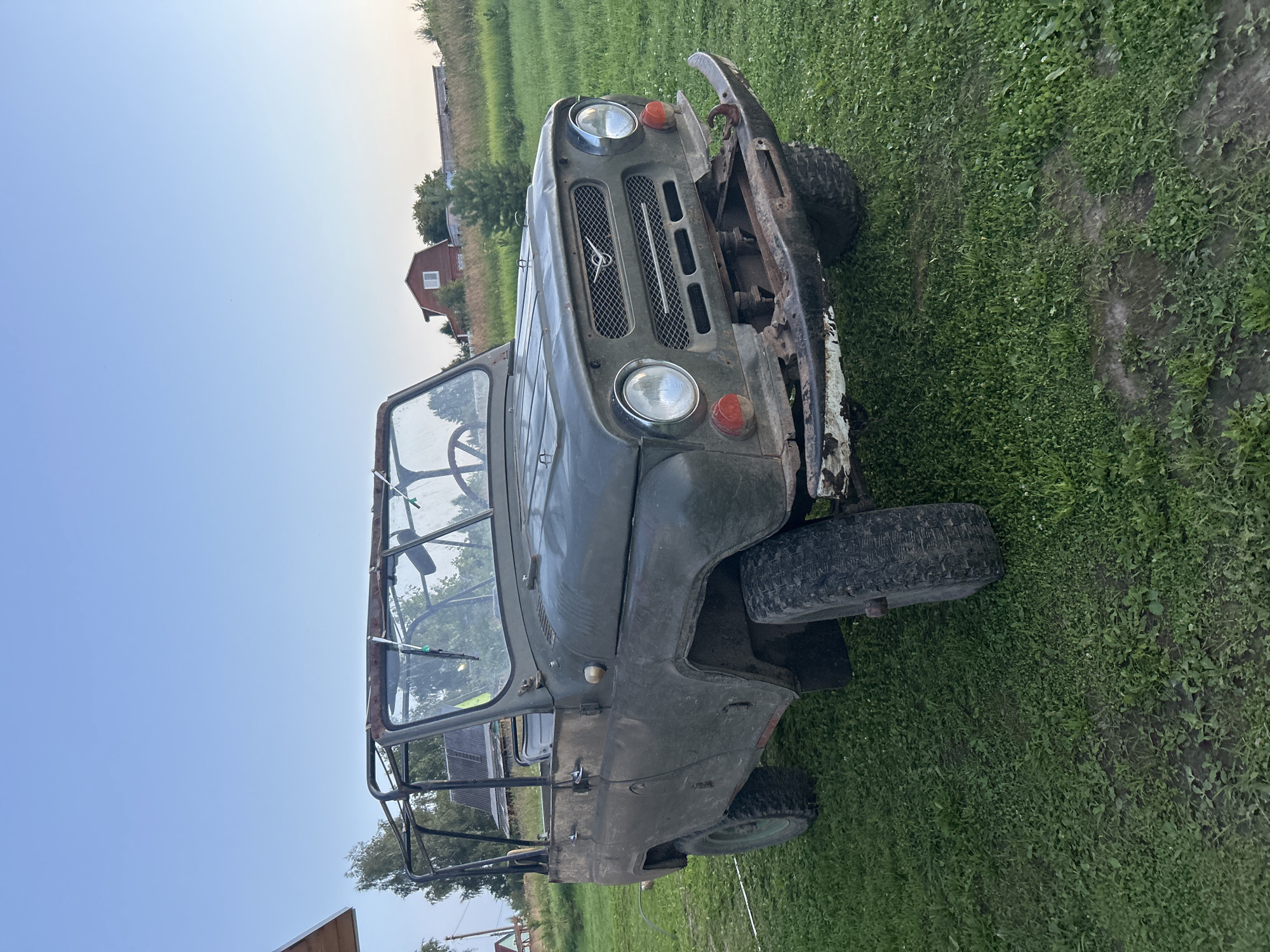 An olive-drab, open-top, vintage UAZ-469 jeep is parked on a grassy area in front of a rural house. The vehicle appears weathered and possibly abandoned, with rust visible on the roll cage and a dented front bumper. The jeep has its doors removed. In the background, a red house with white trim stands on a gentle slope, surrounded by trees and vegetation. The sky is clear and pale blue, indicating either early morning or late evening. The overall scene suggests a quiet, rural setting, likely in the village of Vereiki, Russia, given the architectural style and the prevalence of UAZ vehicles in the region.
