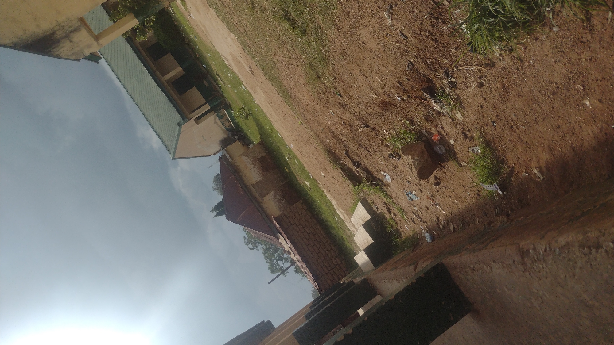 This is an outdoor, eye-level, wide-angle photograph of a semi-developed area, likely in Jos, Nigeria. The image is oriented sideways.
The foreground is dominated by a textured, reddish-brown dirt path with scattered patches of green grass and some litter. A concrete structure, possibly a wall or raised platform, occupies the lower right portion of the frame, casting a shadow. In the mid-ground, a building with a green-tiled roof and light-colored walls is visible. It appears to be a school or institutional building, with visible windows and architectural details. Adjacent to this building, another structure with a red-tiled roof and brick walls is present, suggesting residential or administrative use.
The background features a bright, overcast sky, indicating it is daytime. The overall scene depicts an outdoor environment that is a mix of constructed buildings and uncultivated land, with evidence of human activity in the form of litter on the ground. There are no people visible in the image. The lighting suggests it is daytime, and the overcast sky implies potentially diffused light. The presence of both buildings and the dirt path with grass and litter suggests a location that is not a pristine natural environment but also not a densely urbanized area.