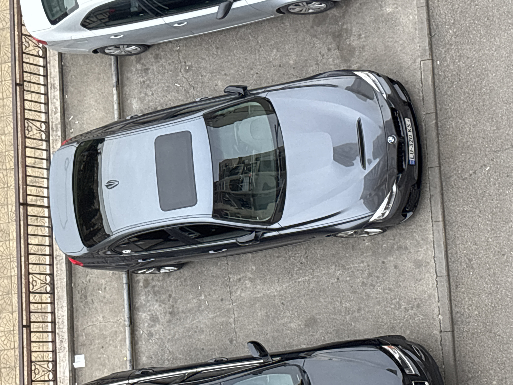 The image shows a view from above onto a street in Tbilisi, Georgia. Three parked cars are visible. At the top is a light gray car, followed by a dark gray car with a sunroof and the license plate "LU-320-KS". The bottom of the image features the front of a black car. A section of a metal fence is seen on the left side. The street surface is concrete. There are no people visible in the image.