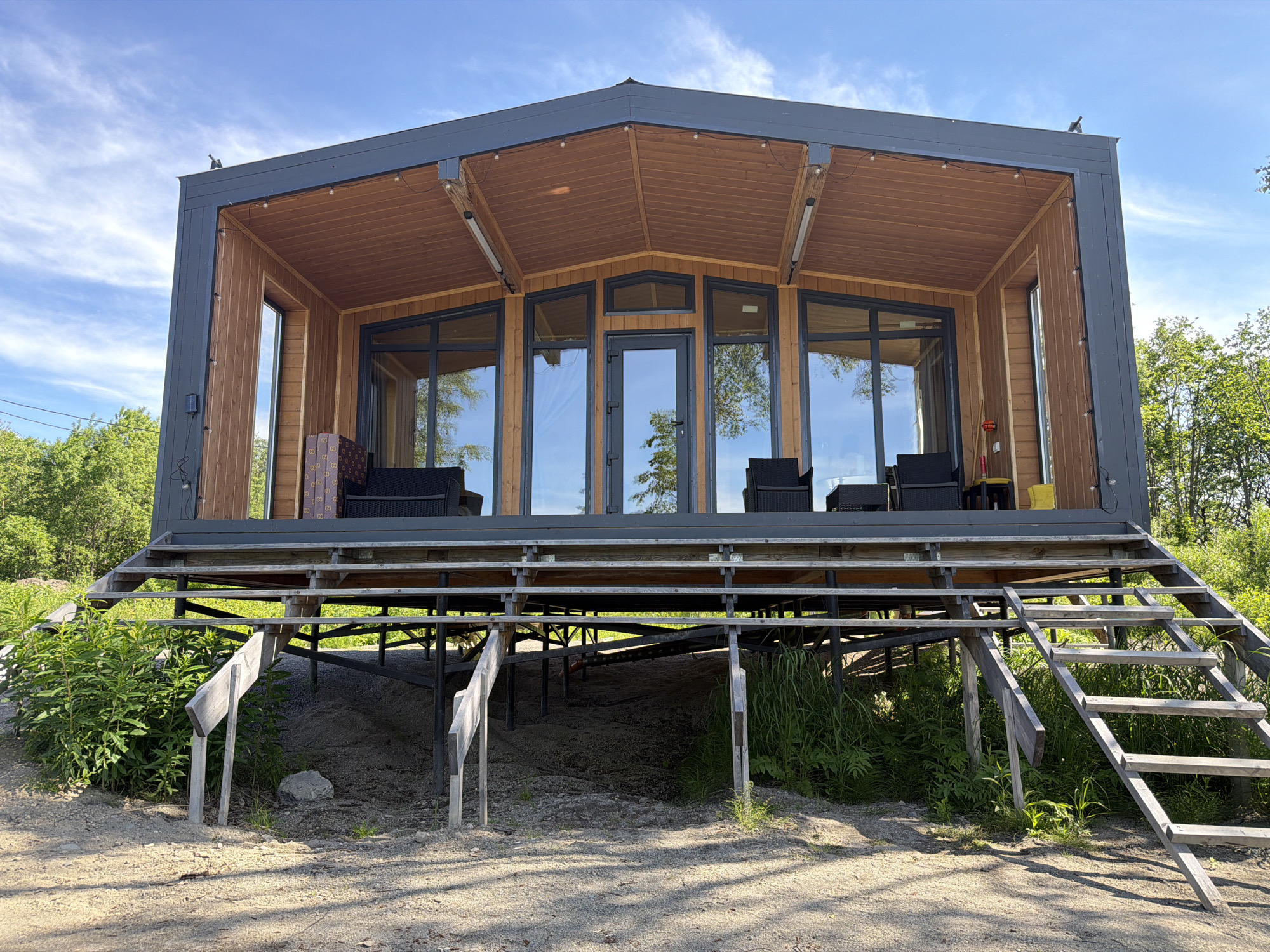 The image depicts a modern, elevated wooden and metal house with large glass windows and doors, situated on a raised wooden deck supported by metal stilts. The house is surrounded by lush green trees and vegetation. A wooden staircase leads up to the deck from the ground, which is covered in gravel and sand. The sky above is a clear blue with a few wispy clouds. The overall scene suggests a rural or semi-rural setting, possibly a vacation home or a modern cabin. The house appears to be in good condition, with string lights visible along the overhang of the deck. There are no people visible in the image. The specific location in the city of Новый, Russia, is not discernible from the visual information provided.