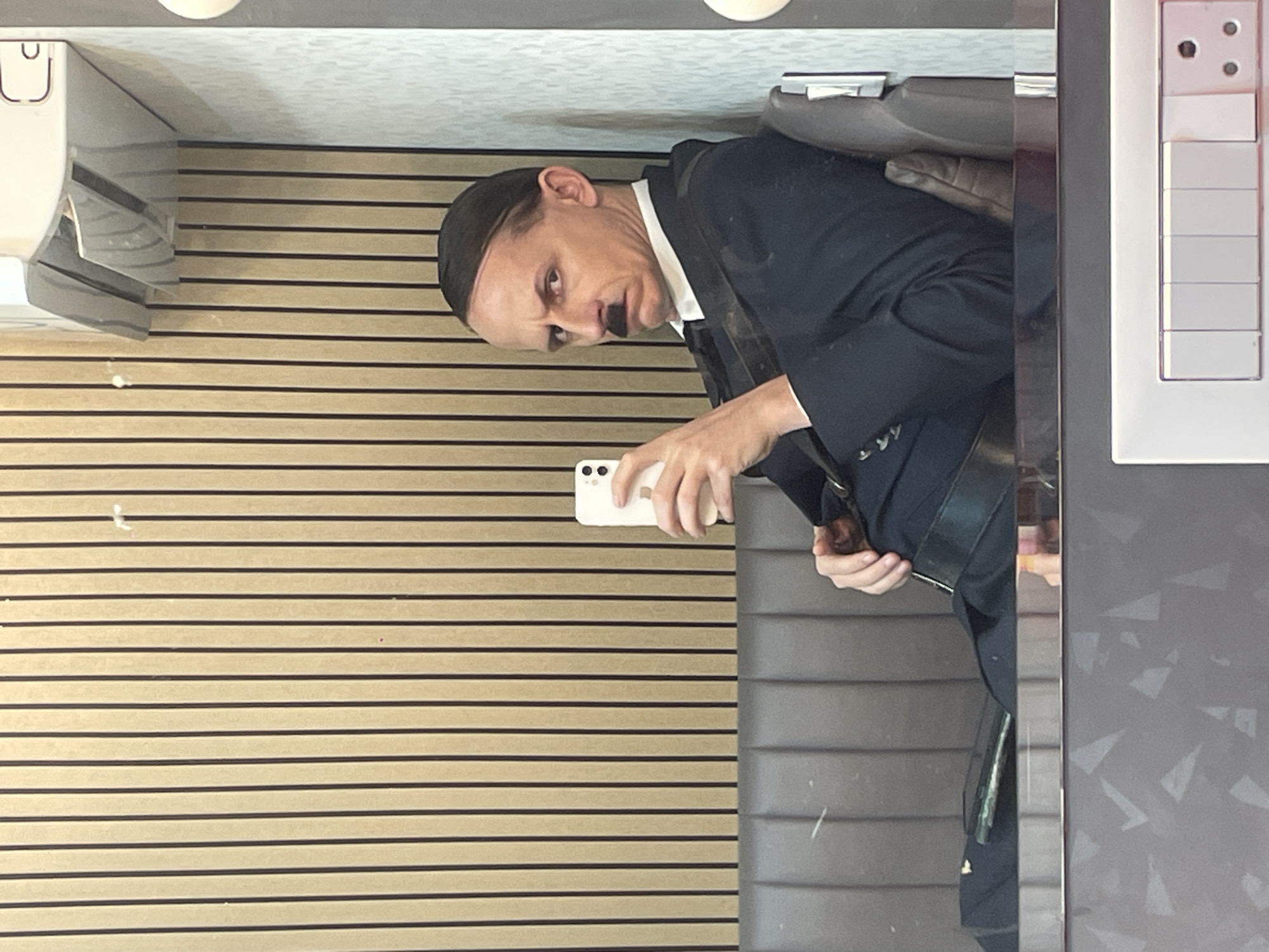 A person dressed as Adolf Hitler, complete with a toothbrush mustache and characteristic hairstyle, sits on a cushioned bench and takes a selfie with a white smartphone. The person is wearing a dark suit, white shirt, and black tie. The background consists of vertical wooden paneling and a gray textured wall. To the right, a white electrical outlet with multiple switches is visible. A reflection in what appears to be a mirror or glass surface shows a portion of a gray seating area with patterned wallpaper below. The scene is indoors.