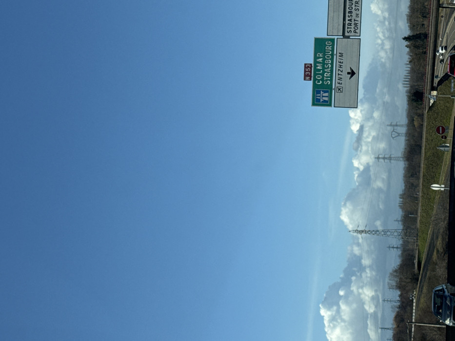 The image depicts a multi-lane highway scene under a clear blue sky with scattered white cumulus clouds along the horizon. In the foreground, traffic moves on the highway, with at least two vehicles visible: a dark gray sedan in the lower left moving left-to-right, and a red vehicle in the lower right, also moving left-to-right. A prominent green and white overhead road sign on the right directs traffic via route N 353 towards COLMAR and STRASBOURG, with a separate white panel indicating an exit for ENTZHEIM. A partial sign to the right also mentions STRASBOURG and PORT DE STRASBOURG. These locations are in France, specifically near Strasbourg in the Alsace region. The midground features a landscape with bare deciduous trees and numerous tall metal lattice power line towers stretching across the horizon. The perspective suggests the photograph was taken from a vehicle in motion.