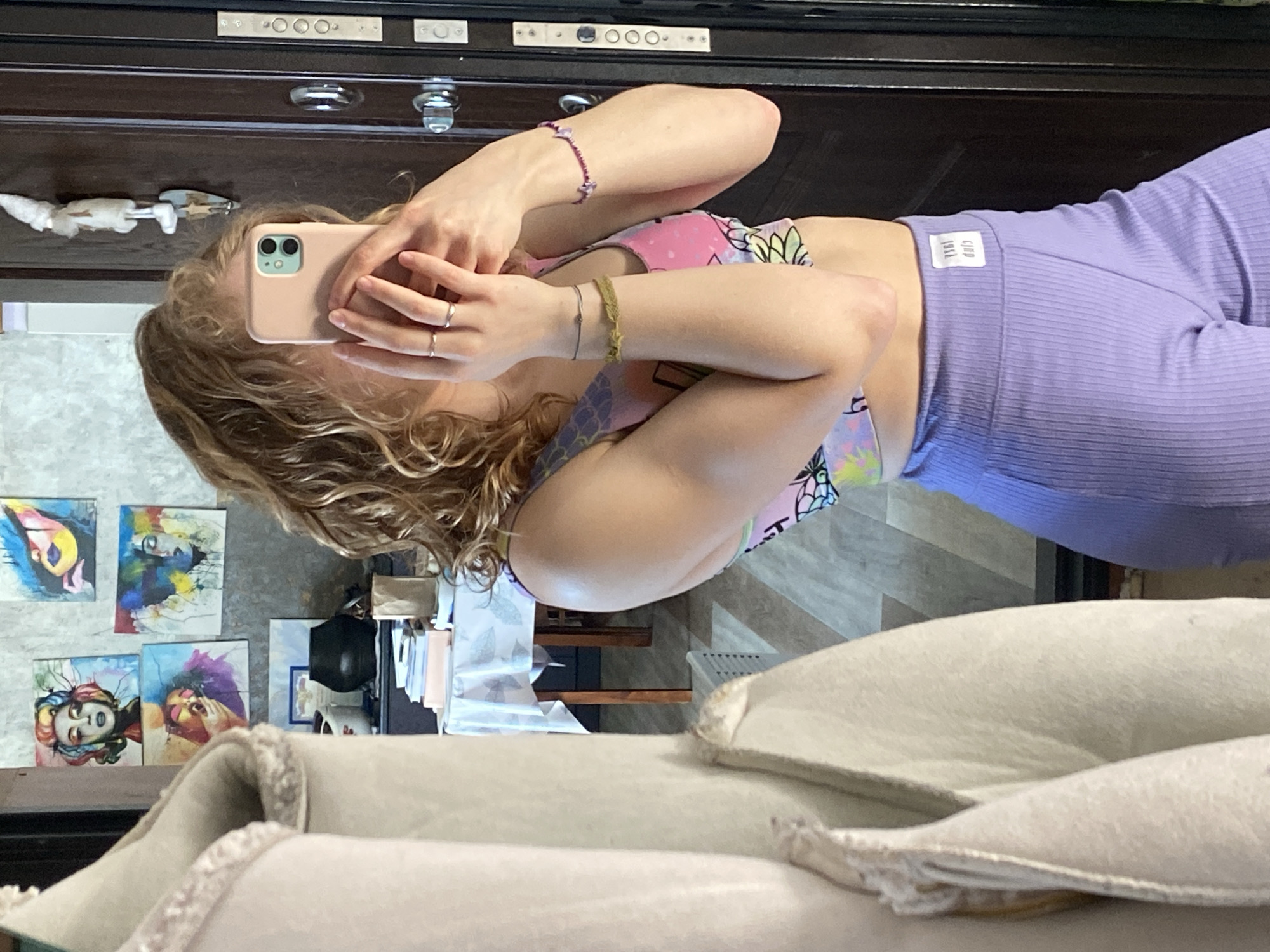 A woman is taking a selfie in front of a mirror. She is wearing a sports bra with a pineapple pattern and light purple ribbed leggings. She holds a pink-cased iPhone 12 or 13 in her hands. She is wearing multiple bracelets and rings. The background includes a dark wooden dresser with silver hardware. A wall is visible with several colorful art pieces. A white sofa with decorative trim is in the foreground. It is not possible to determine the location in the city of Chaikovsky, Russia from the image.