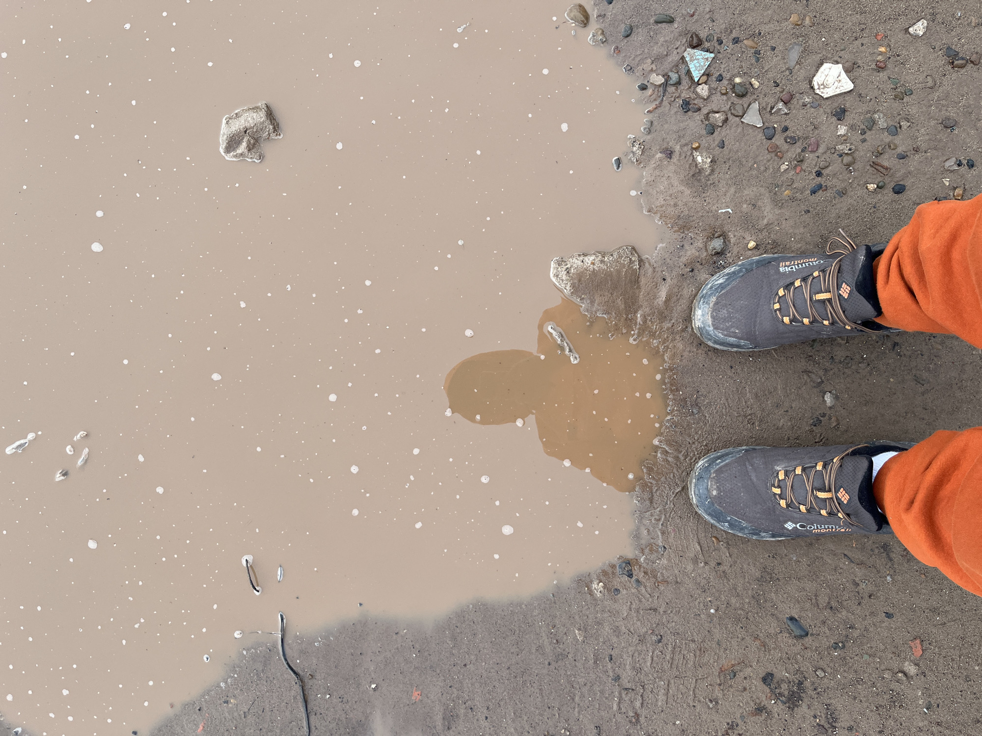 A top-down shot depicts a mud puddle with a pair of Columbia Montrail hiking boots and orange pants visible on the right side. The puddle is a murky brown color with white splatter marks. Debris like rocks and small pieces of trash are visible around the edge of the puddle. The ground surrounding the puddle appears to be a mix of dirt, mud, and small pebbles. The location cannot be determined from the image.