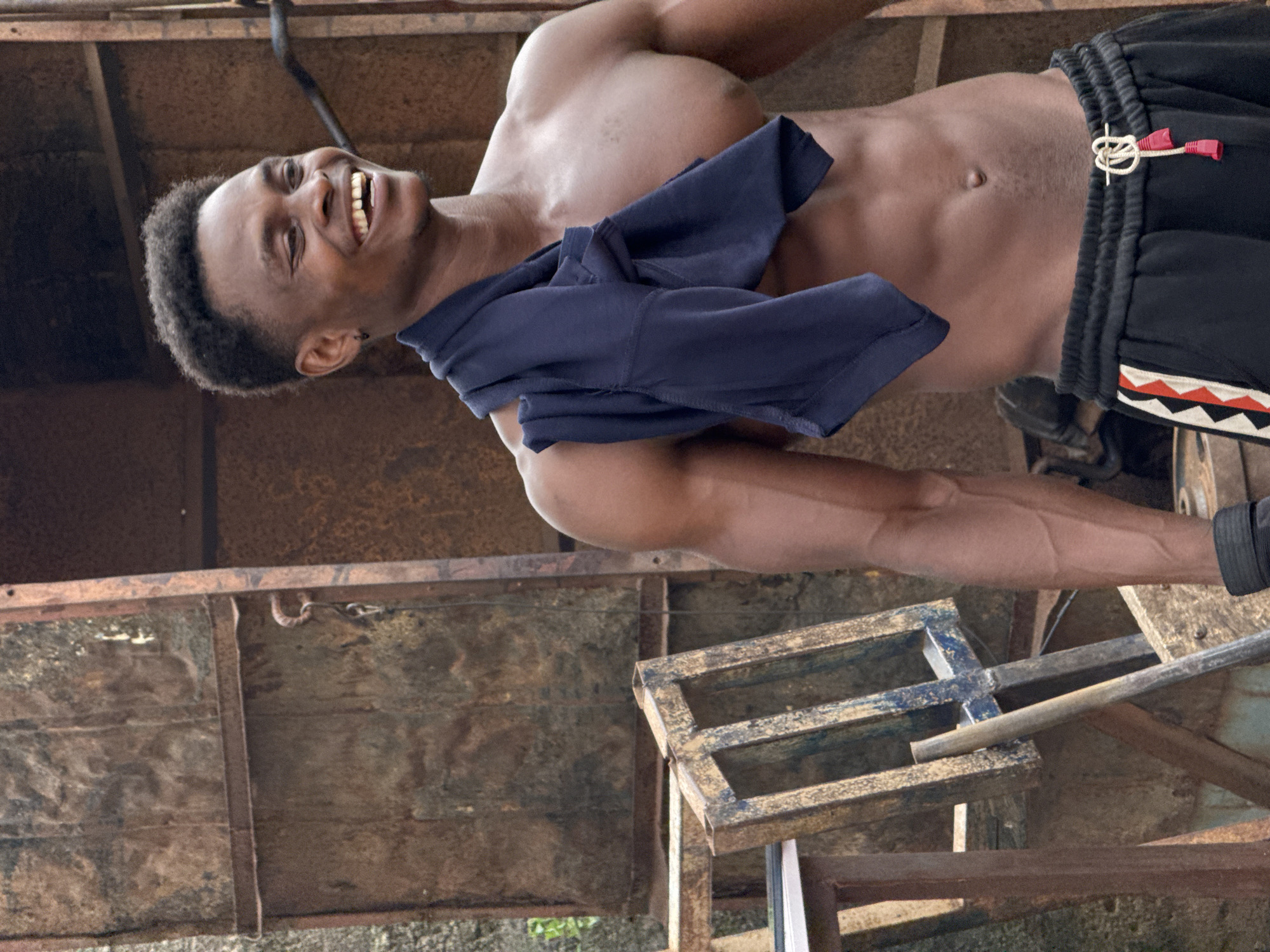 A shirtless Black male with short, dark, curly hair and an earring smiles broadly while standing in an outdoor urban environment. He has a dark blue shirt draped over his shoulders and is wearing black athletic shorts with red and white striped accents on the side. His muscular physique is visible, with well-defined abs and arms. The background consists of weathered, rusty metal structures and some wooden elements, suggesting a potentially industrial or junkyard setting. The lighting suggests it is daytime. The location is identified as Akwanga, Nigeria.