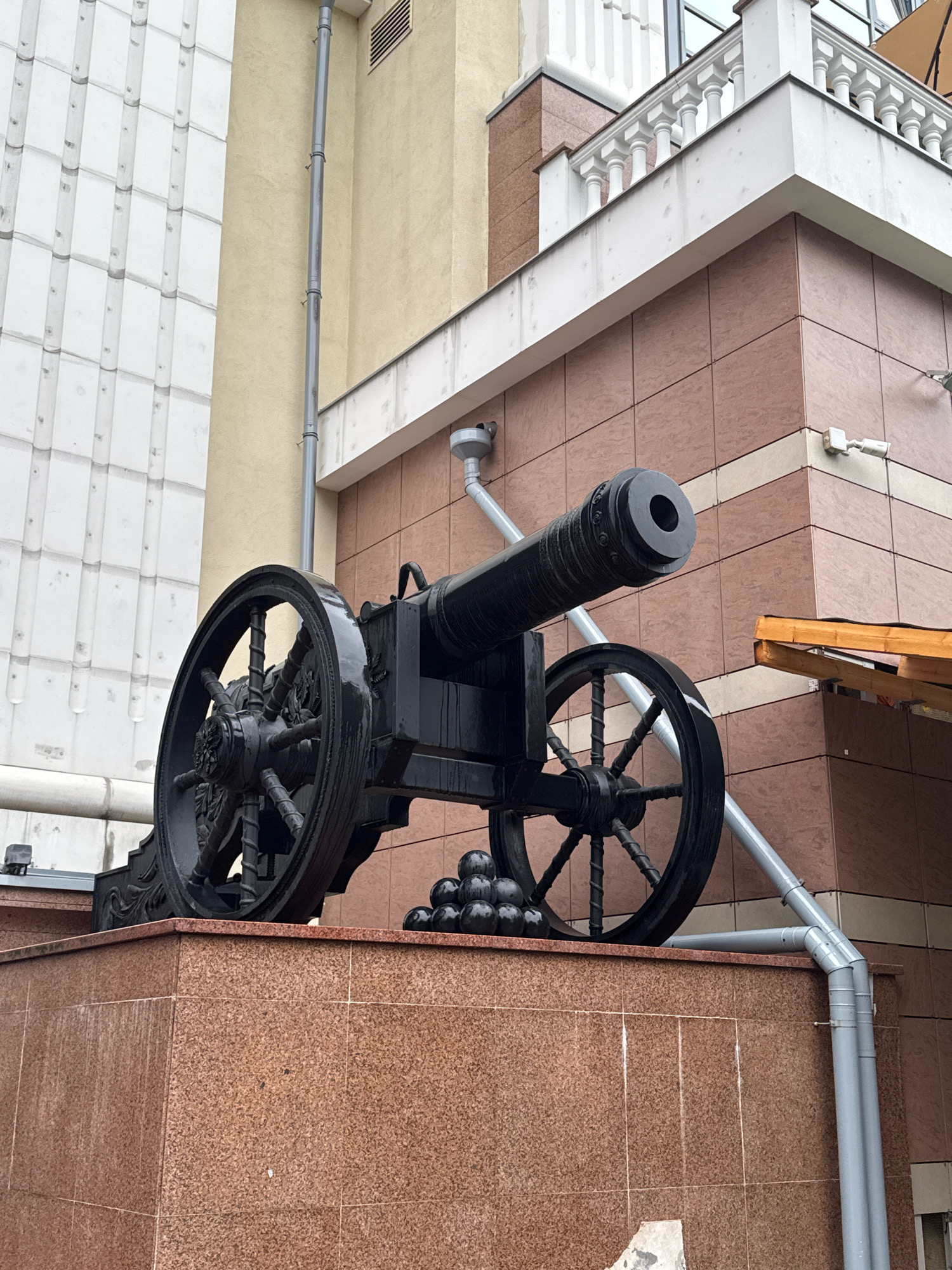 The image depicts an antique black cannon displayed on a granite pedestal outside a building in Saint Petersburg, Russia. The cannon is positioned on a wheeled carriage with decorative spokes. A pile of cannonballs is visible in front of the cannon. The building behind the cannon has a modern facade with tiled walls in shades of pink and beige, and white architectural elements. The scene appears to be outdoors, with a downspout running along the side of the building. There are no people visible in the image.