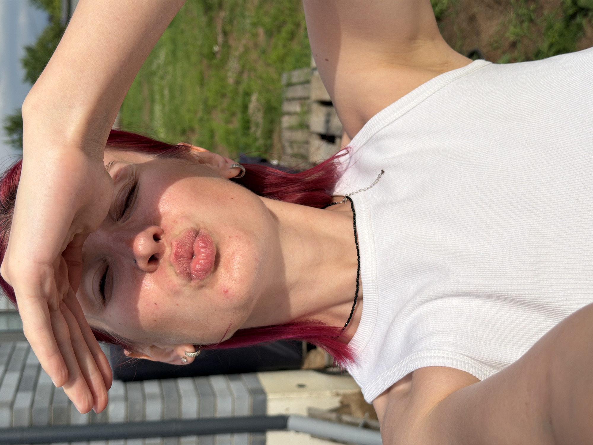 This is a close-up, vertically oriented selfie of a young person with vibrant red hair, taken outdoors during the daytime. The subject is wearing a white ribbed t-shirt and a black beaded necklace. Their right hand is raised, shielding their eyes from the sun, and their left eye is closed. Their lips are puckered, suggesting they are making a kissing face. They have a nose ring and a subtle earring in their left ear. The background is slightly blurred, showing green grass and some wooden structures, possibly indicating a backyard or park setting. The lighting suggests it is a sunny day. The image is captured from a high angle, looking down at the subject. No visible text is present.