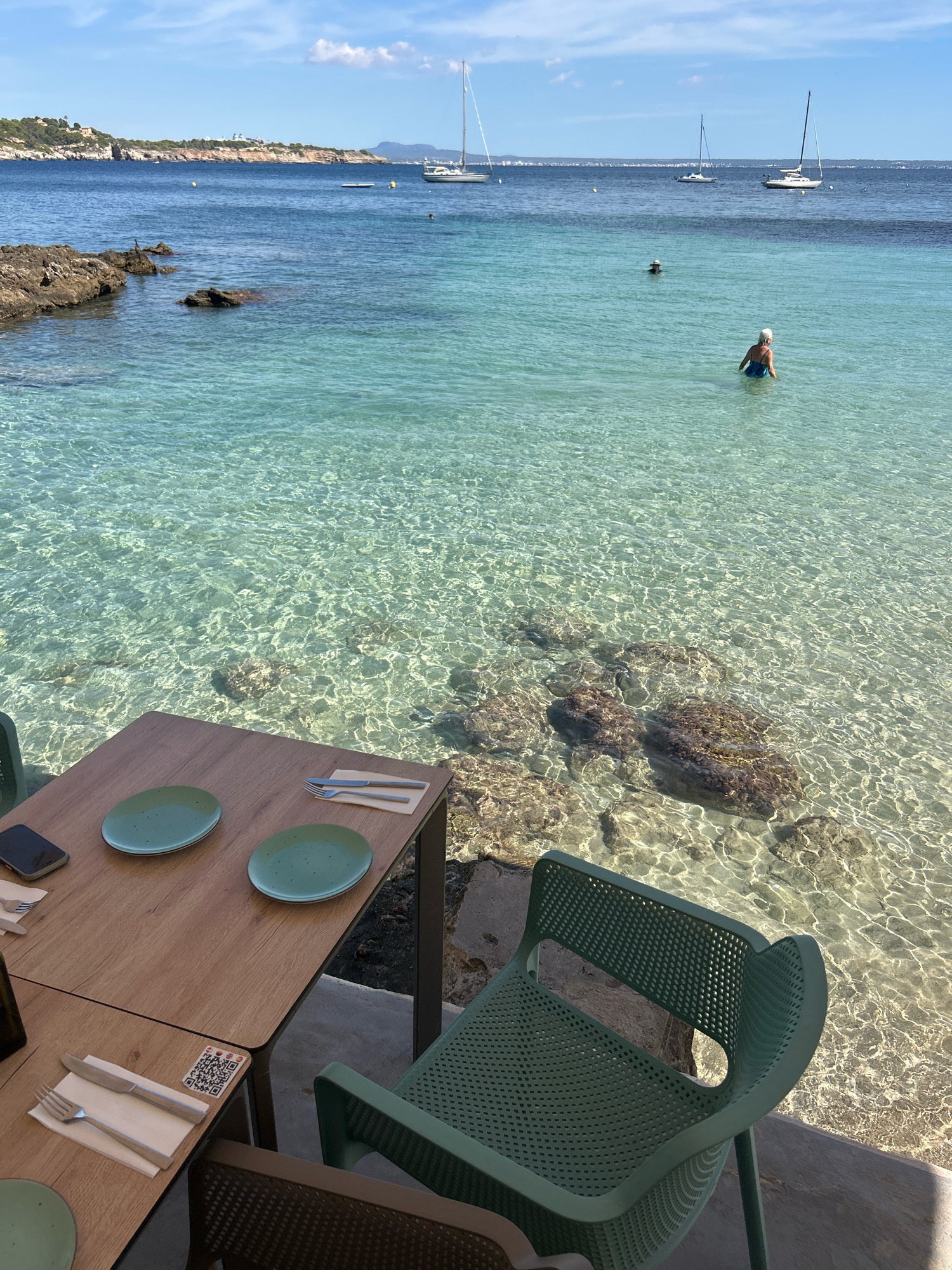 In the city of Cas Català-ses Illetes, Spain, a table is set for two with plates and cutlery by the crystal-clear waters of the Mediterranean Sea. The sun casts dappled patterns on the sandy seabed, visible through the transparent water. Two green plastic chairs flank the table, with another chair partially visible in the foreground. In the background, sailboats dot the horizon, and a few people are enjoying a swim in the calm sea. The scene evokes a sense of tranquil seaside dining.