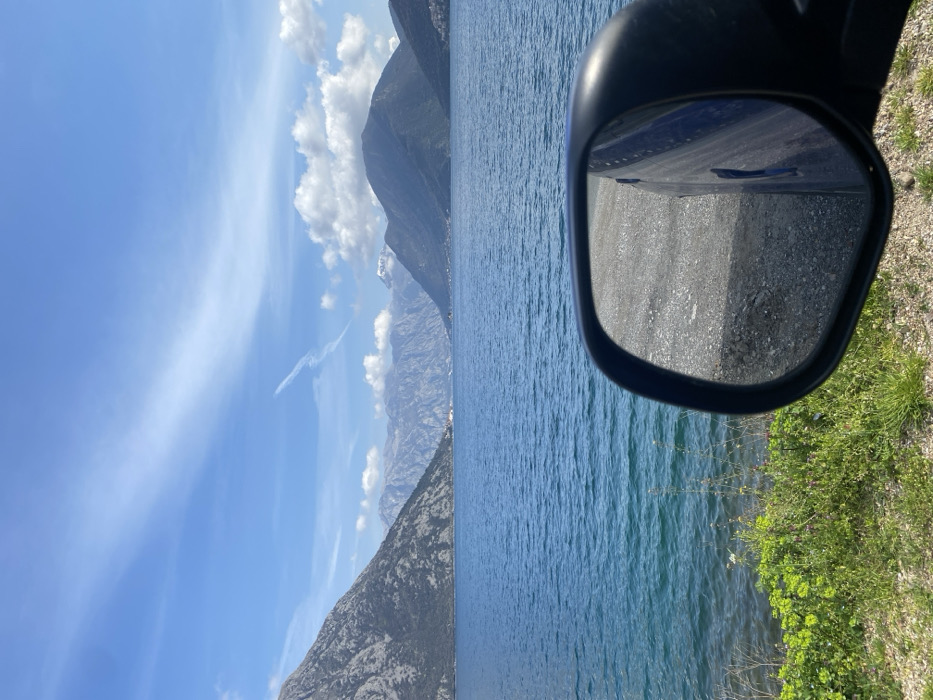 The image displays a panoramic view of the Bay of Kotor from a roadside vantage point in Lipci, Montenegro. The scene is dominated by a large expanse of vibrant blue water, showing subtle surface ripples, extending towards a distant mountainous landscape under a bright blue sky with scattered white clouds.
In the lower right foreground, a black plastic vehicle side mirror is prominently featured, its reflective surface showing a close-up of a light gray, gravelly ground surface and a portion of a dark blue vehicle body. Along the immediate left edge of the water, a narrow strip of green and brown earth is visible, supporting patches of bright green foliage and sparse purple and white wildflowers.
The midground features the extensive blue waters of the bay, which are framed by steep, verdant and rocky mountains that rise directly from the water's edge on both sides. In the far distance, additional layers of mountains appear, some with visible grey rock formations and one distinct peak displaying a patch of white, likely snow. Small clusters of buildings or settlements are discernible along the distant shorelines at the bases of these mountains.
The sky occupies the upper portion of the image, presenting a clear blue with several distinct white cumulus clouds, primarily on the right side. A long, thin white contrail or wispy cloud formation stretches diagonally across the upper-mid sky. No individuals or direct actions are depicted in the static scene.