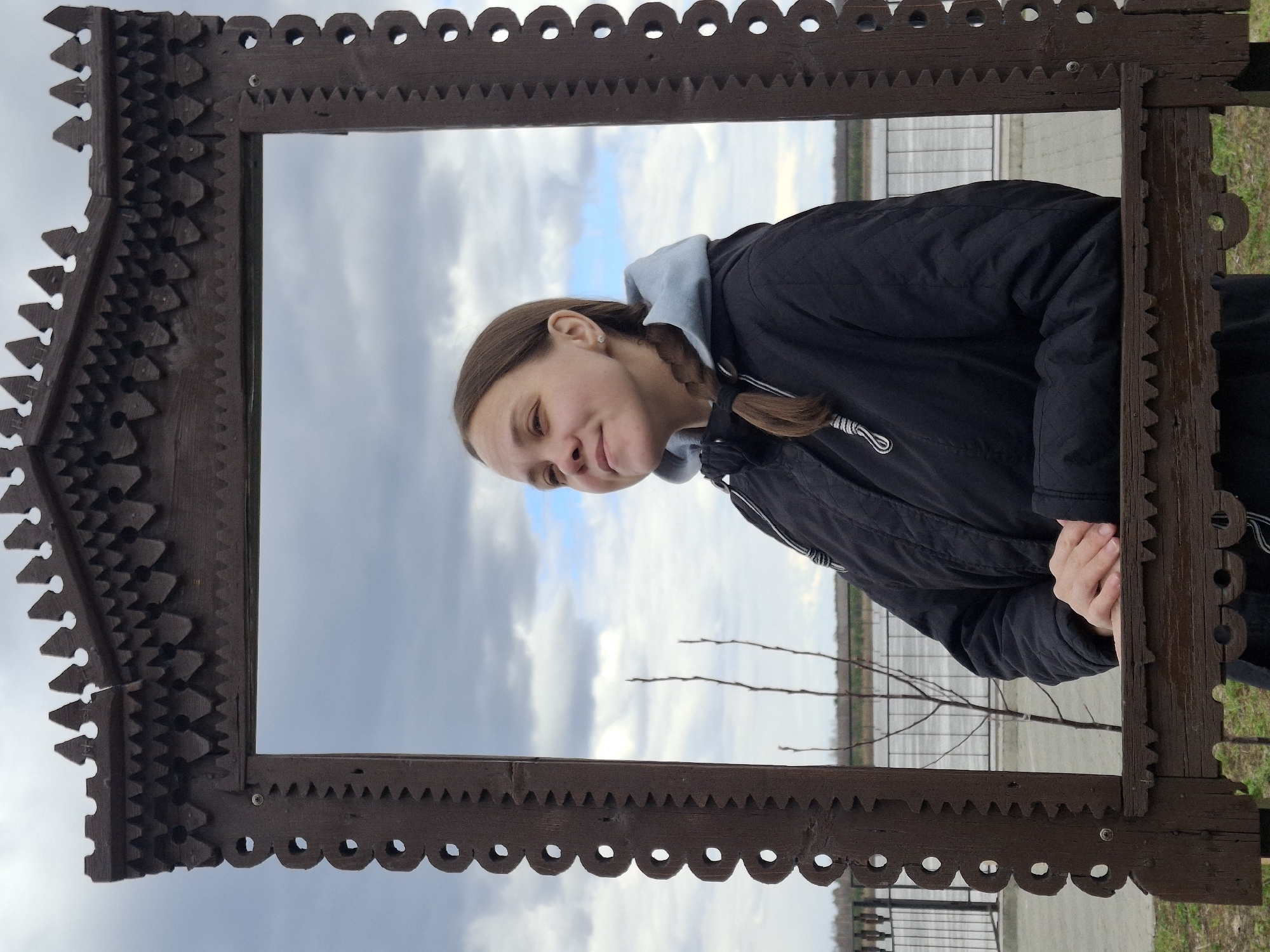 The image features a young woman leaning into a large, ornate wooden frame, posing for a photograph. The frame is intricately carved with decorative patterns, including scalloped edges and sawtooth designs, and appears to be made of dark, possibly weathered wood. The woman, with brown hair tied in a braid, is wearing a black jacket over a light blue hooded sweatshirt. She looks towards the viewer with a gentle, slight smile, conveying a relaxed and pleasant demeanor. Her hands are visible, resting on the lower part of the frame.
Through the opening of the frame, a wide body of water, likely a river or lake, stretches into the background under a predominantly cloudy sky. Patches of lighter blue are visible amidst the grey clouds. A metal fence or railing runs along the distant waterfront, and a few bare tree branches are also discernible. The setting suggests an outdoor location, possibly a river embankment or park. The overcast conditions indicate it is daytime. This scene is consistent with the provided location context of Sarapul, Russia, which is situated on the Kama River. There is no visible text in the image.