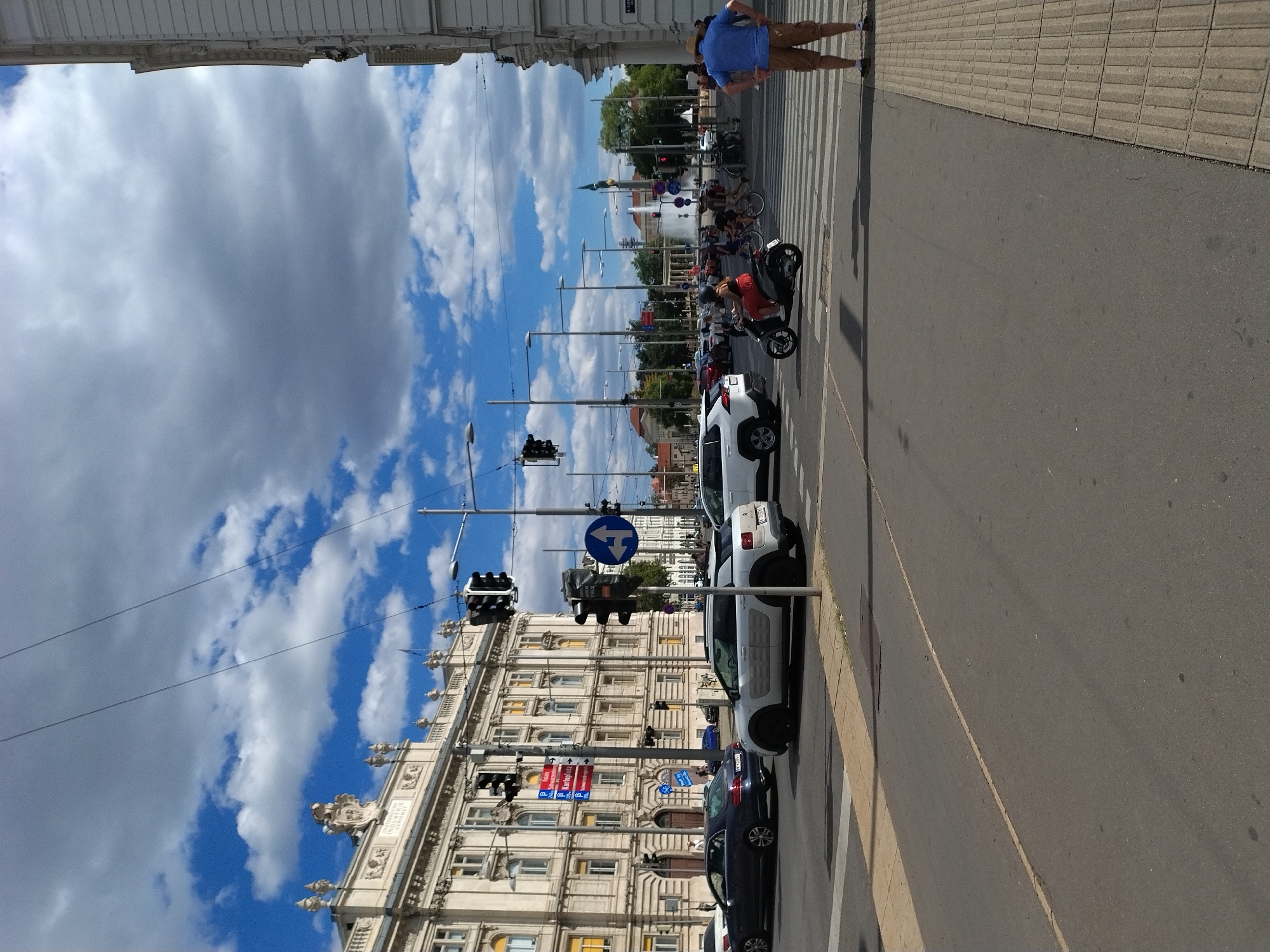 The image shows a street intersection in Vienna, Austria, on a sunny day with scattered clouds. In the foreground, a grey asphalt road dominates the lower half of the frame, with a yellow line marking a lane. A pedestrian crossing with white stripes is visible. To the right, a light-colored tiled sidewalk is present.
On the sidewalk, a man wearing a blue t-shirt and brown shorts stands with his hands on his hips, facing away from the camera.
In the middle ground, several vehicles are present. A white Citroën C4 Cactus is stopped at the intersection. To its left, a dark-colored sedan is in motion, and behind it, another white SUV is partially visible. A woman is riding a scooter in the middle of the intersection.
In the background, a large, ornate, light-colored building with many windows and classical architectural details lines the street. Traffic lights and street lamps are visible throughout the intersection. Further in the distance, a large fountain with water spouting upwards is a prominent feature. Trees and other buildings are also visible, suggesting a busy urban environment.