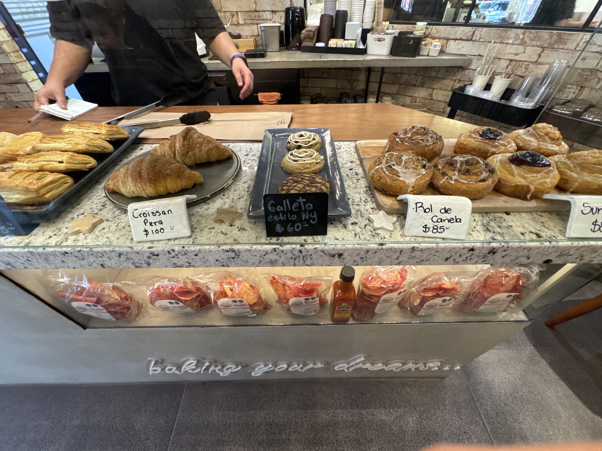 This media file captures the interior of a cafe or bakery, likely in Puebla, Mexico. The scene is dominated by a display counter laden with freshly baked goods, with a person working behind it.
In the foreground, a white speckled counter holds various pastries. To the left, a tray of golden-brown palmiers or similar layered pastries is visible. Next to it, a dark plate displays two sugar-dusted croissants, with a small sign reading "Croissan Pera $100" (Pear Croissant). In the center, a tray presents three cookies, two with white icing and one with chocolate drizzle, labeled "Galleta estilo Ny $60" (New York style cookie). To the right, a wooden board holds several cinnamon rolls, some topped with white icing and others with blueberries, accompanied by a sign that says "Rol de Canela $85" (Cinnamon Roll). Another partially visible sign reads "Sur $8," likely indicating another pastry.
Behind the counter, a person, visible from the chest up, wears a black shirt and has their hands near a white notepad and pen, possibly taking an order or preparing paperwork. Tongs are also on the wooden counter surface. The background features a brick-patterned wall, with shelves holding various coffee supplies like disposable cups, stirrers, and a coffee machine.
Below the main counter, a glass display case reveals several clear plastic bags of packaged snacks labeled "MALANGA," appearing to be root vegetable chips. A bottle of hot sauce is also placed among them. The lower part of the display case features raised white lettering that spells out "baking your dreams."
The overall setting suggests a bustling, inviting bakery or coffee shop, with a focus on fresh pastries and a modern, clean aesthetic. The prices are consistent with Mexican Pesos, aligning with the Puebla, Mexico location context.