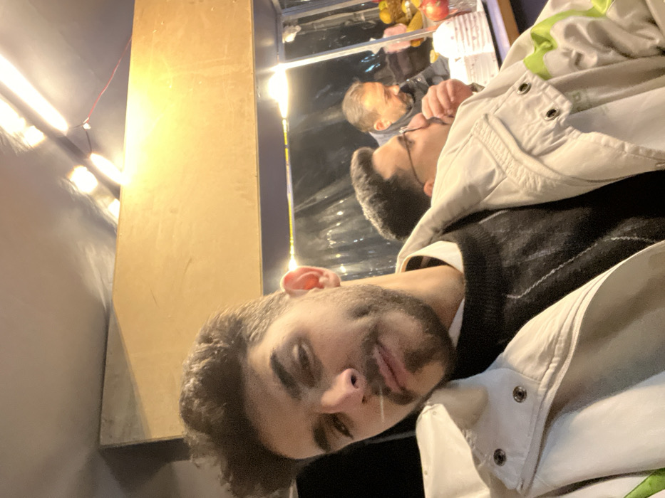 A man with dark hair and a beard is looking at the camera. He is wearing a vest and a light-colored jacket. Behind him is a reflective surface showing another man with glasses eating something. A column is next to the reflective surface and there is a row of lights to the side.