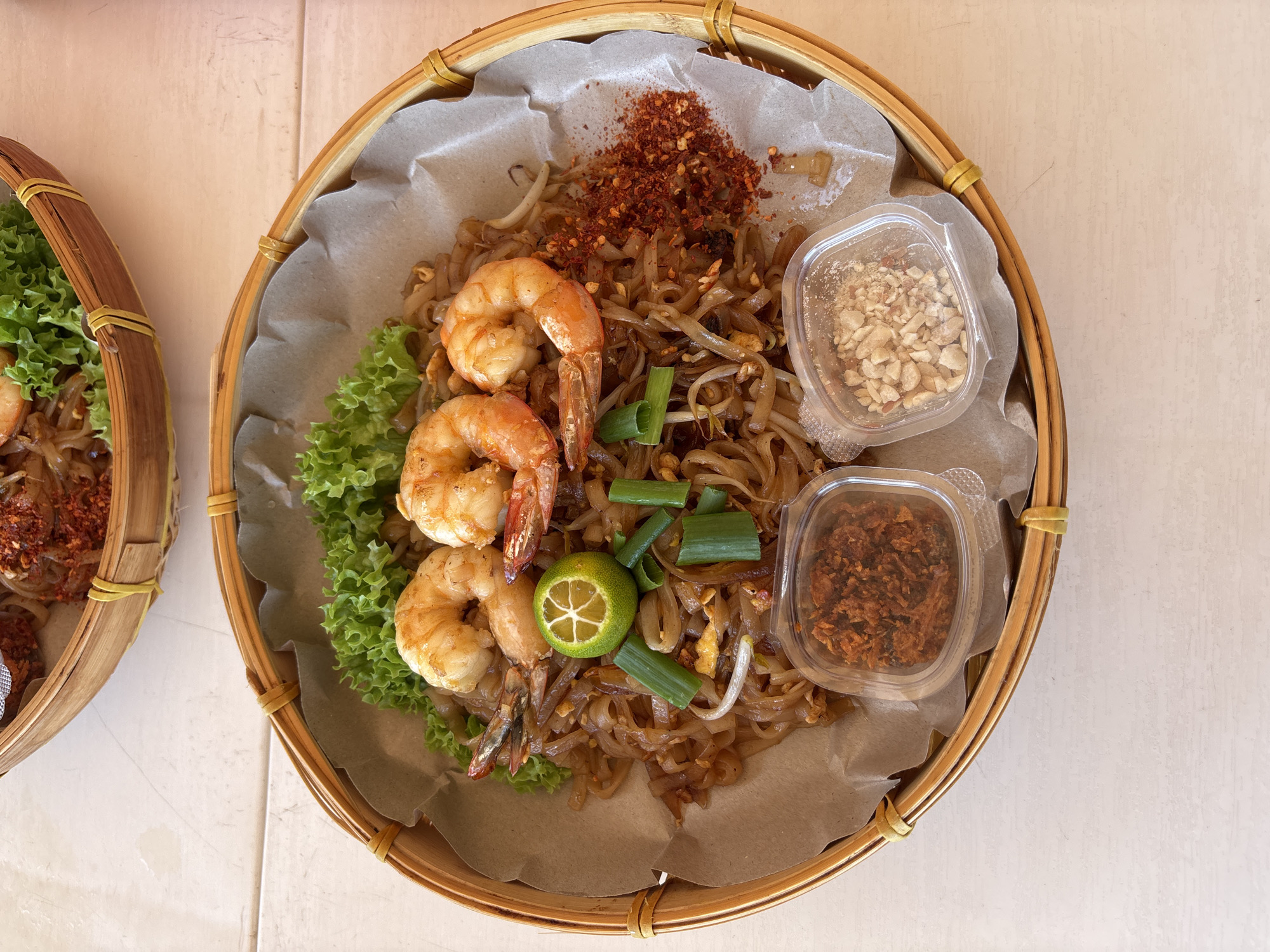 An overhead shot shows a serving of Pad Thai in a woven bamboo basket lined with brown parchment paper. The Pad Thai features rice noodles, bean sprouts, green onions, and scrambled egg, topped with three large, cooked shrimp. A lime wedge, a small pile of red chili flakes, and two small plastic containers filled with crushed peanuts and fried shallots are also present. In the background, another basket of Pad Thai is partially visible. The dishes are placed on a white, wood-grained surface, likely a table. The image appears to be taken at a restaurant in George Town, Malaysia, judging by the style of presentation which is common in the region.