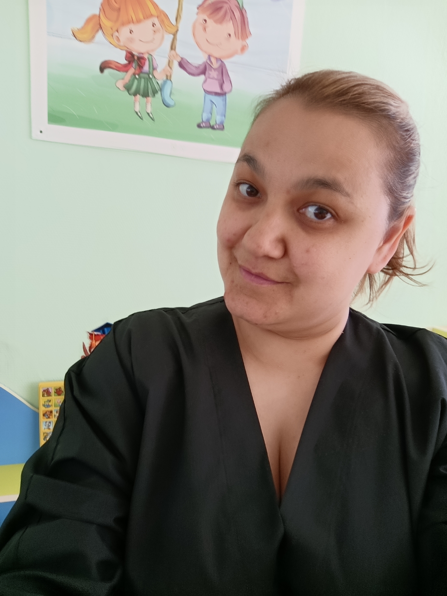 A woman is facing the camera with a slight smile. She has fair skin, dark eyes, and light brown hair pulled back. She is wearing a black V-neck garment. Behind her, there is a light green wall. A framed cartoon image of two children playing on a swing is hanging on the wall. A small portion of what appears to be a children's bookcase or display unit with a yellow box is visible on the left side of the frame. The location is potentially Chaikovsky, Russia.