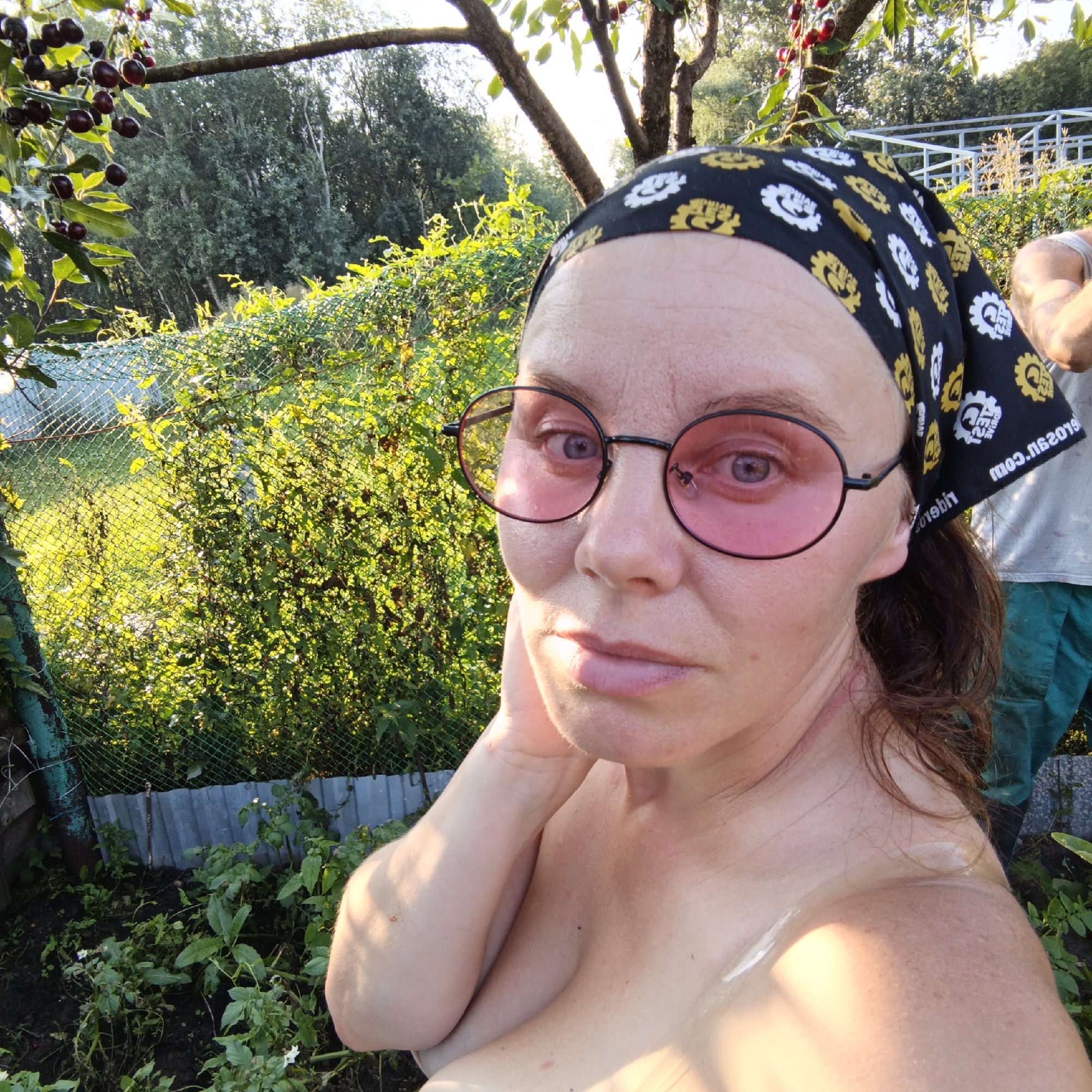 The image is a selfie taken outdoors in a garden or yard setting. A woman with light skin and brown hair is featured in the foreground. She is wearing round, pink-tinted glasses and a black bandana with a yellow and white pattern. Her left arm is bent, and her hand is near her face. The background shows greenery, a fence, and a cherry tree with ripe cherries. The lighting suggests it is daytime, possibly late afternoon due to the warm sunlight. A portion of another person's torso, wearing a grey tank top and green pants, is visible to the right of the woman. The location is specified as Chaikovsky, Russia.