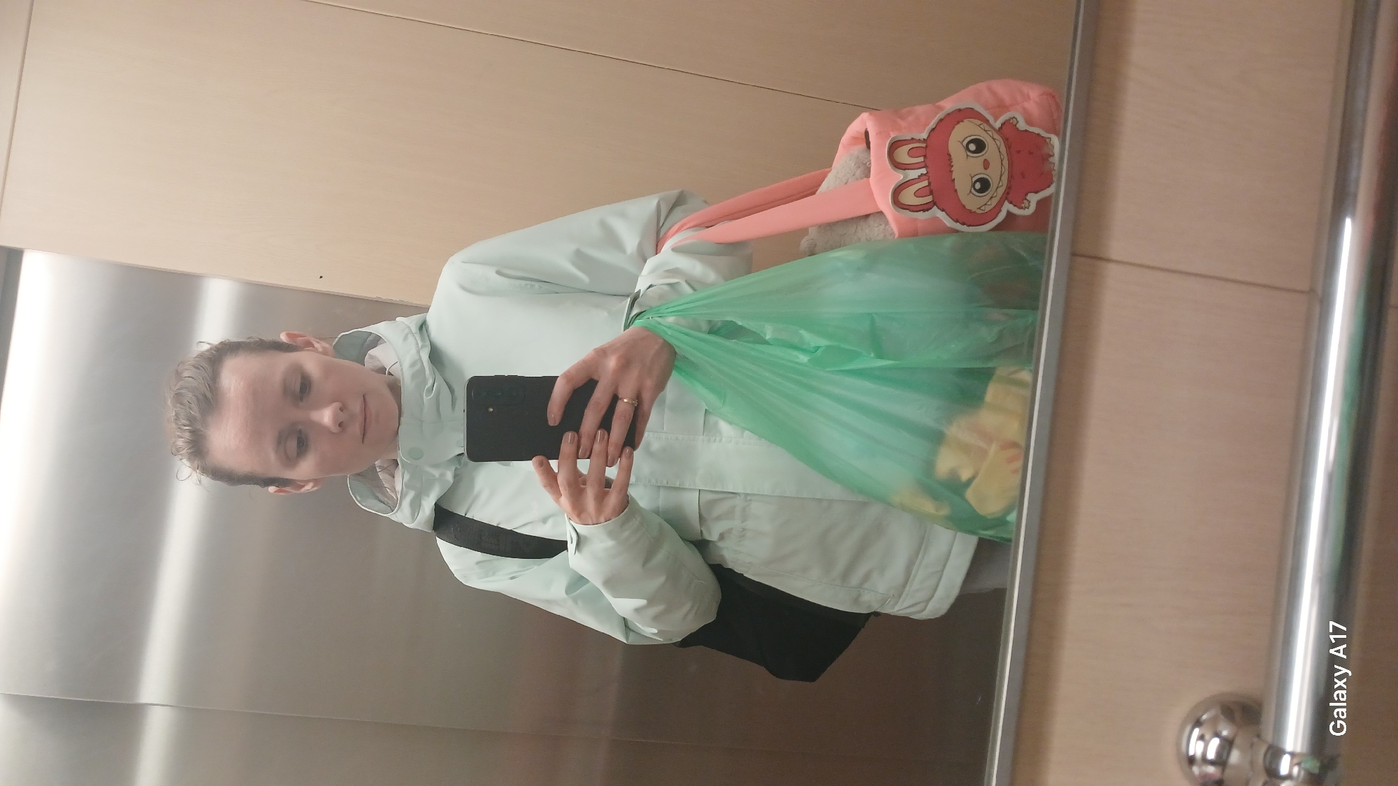 A person is taking a selfie in an elevator. The person is wearing a light blue jacket and a black bag is slung over their shoulder. They are holding a black smartphone. A pink tote bag with a cartoon character and a green plastic bag are also visible. The elevator has stainless steel walls and a wooden panel. "Galaxy A17" is written on the lower right corner of the reflection. The location cannot be determined from the image.