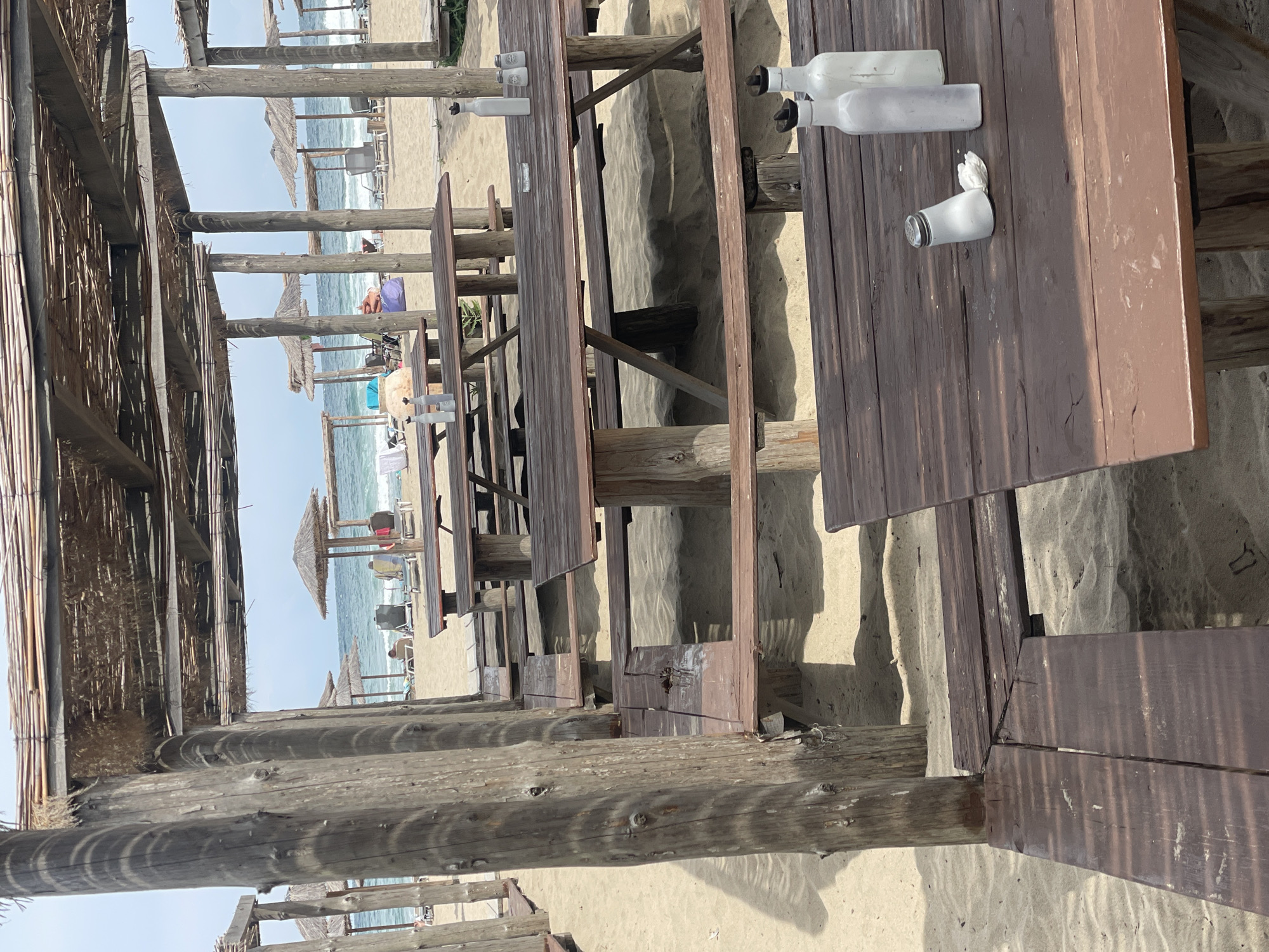 The image depicts a beachside restaurant in Rezovo, Bulgaria. Numerous wooden picnic tables and benches are arranged under thatched-roof pergolas on a sandy beach. In the foreground, a table has two white bottles and a salt shaker. Further back, several other tables and benches are visible, some with empty chairs. Beyond the seating area, beach umbrellas and lounge chairs are scattered on the sand. The sea with gentle waves is in the background, under a cloudy sky. A few people are visible in the distance, relaxing on the beach. The overall scene conveys a sense of a quiet, relaxed atmosphere at a coastal establishment.