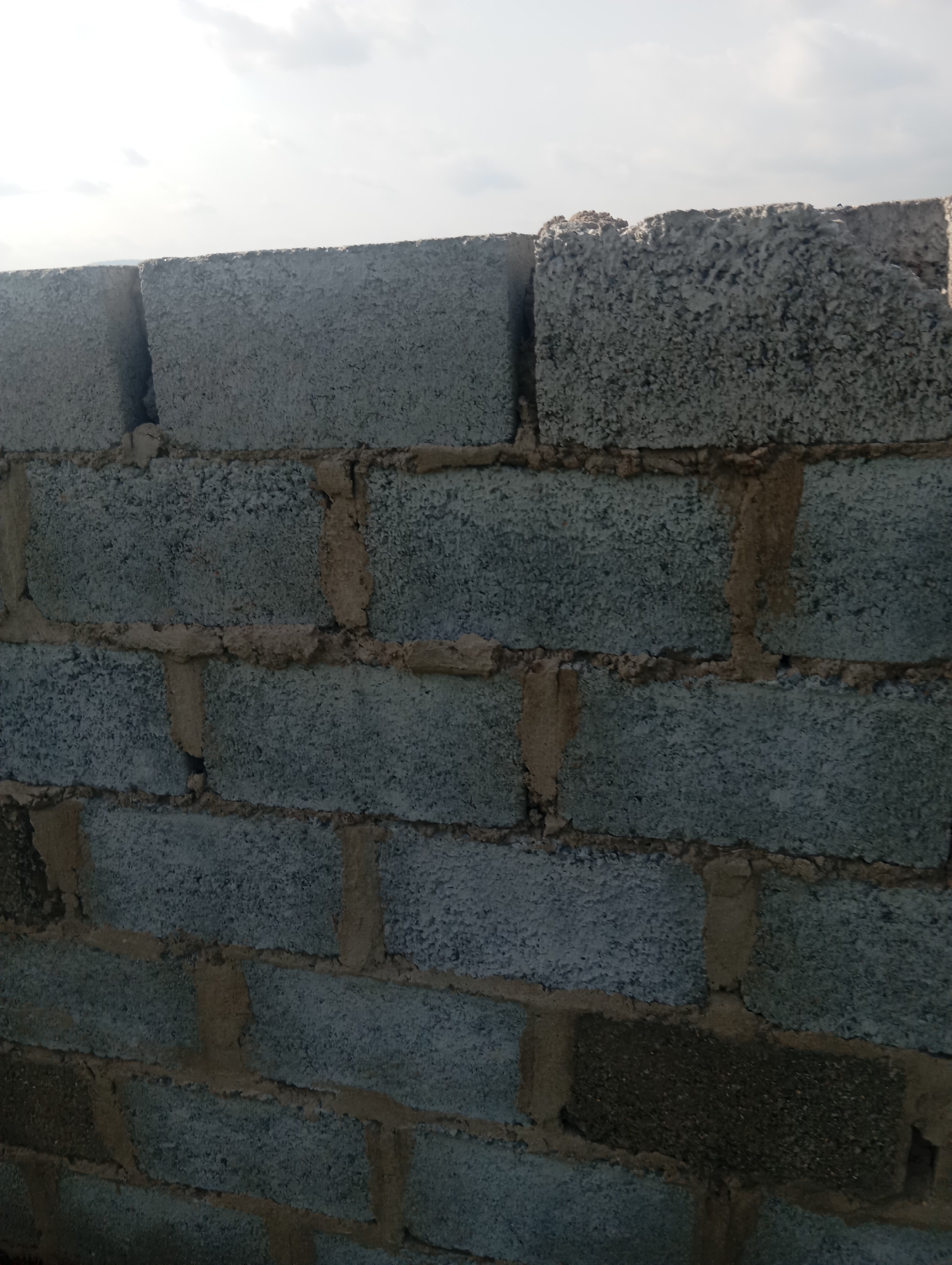 A medium shot captures a wall constructed from grey cinder blocks, laid in horizontal rows with visible tan mortar. The top edge of the wall is uneven, with some blocks broken or protruding. Above the wall, a pale sky with scattered white clouds is visible, suggesting an outdoor setting. The scene appears to be in Dutsen Alhaji, Nigeria.