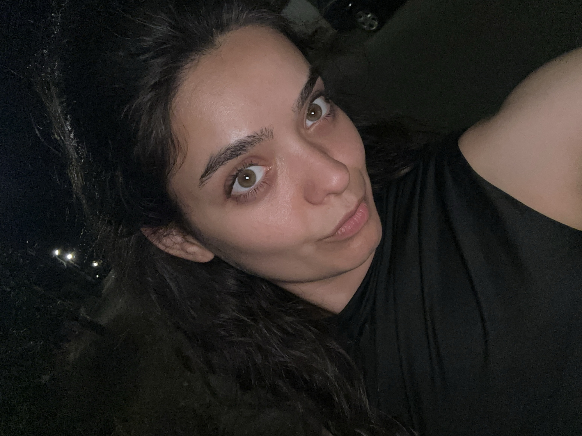 A close-up, nighttime selfie of a young woman with dark, wavy hair and brown eyes. She is wearing a black top and is looking towards the camera. Her skin appears slightly dewy. The background is dark and out of focus, with faint lights visible in the distance, suggesting an outdoor setting. There is a hint of a car on the right side of the image. The specific location in Malevo, Bulgaria, cannot be determined from the visual information provided.