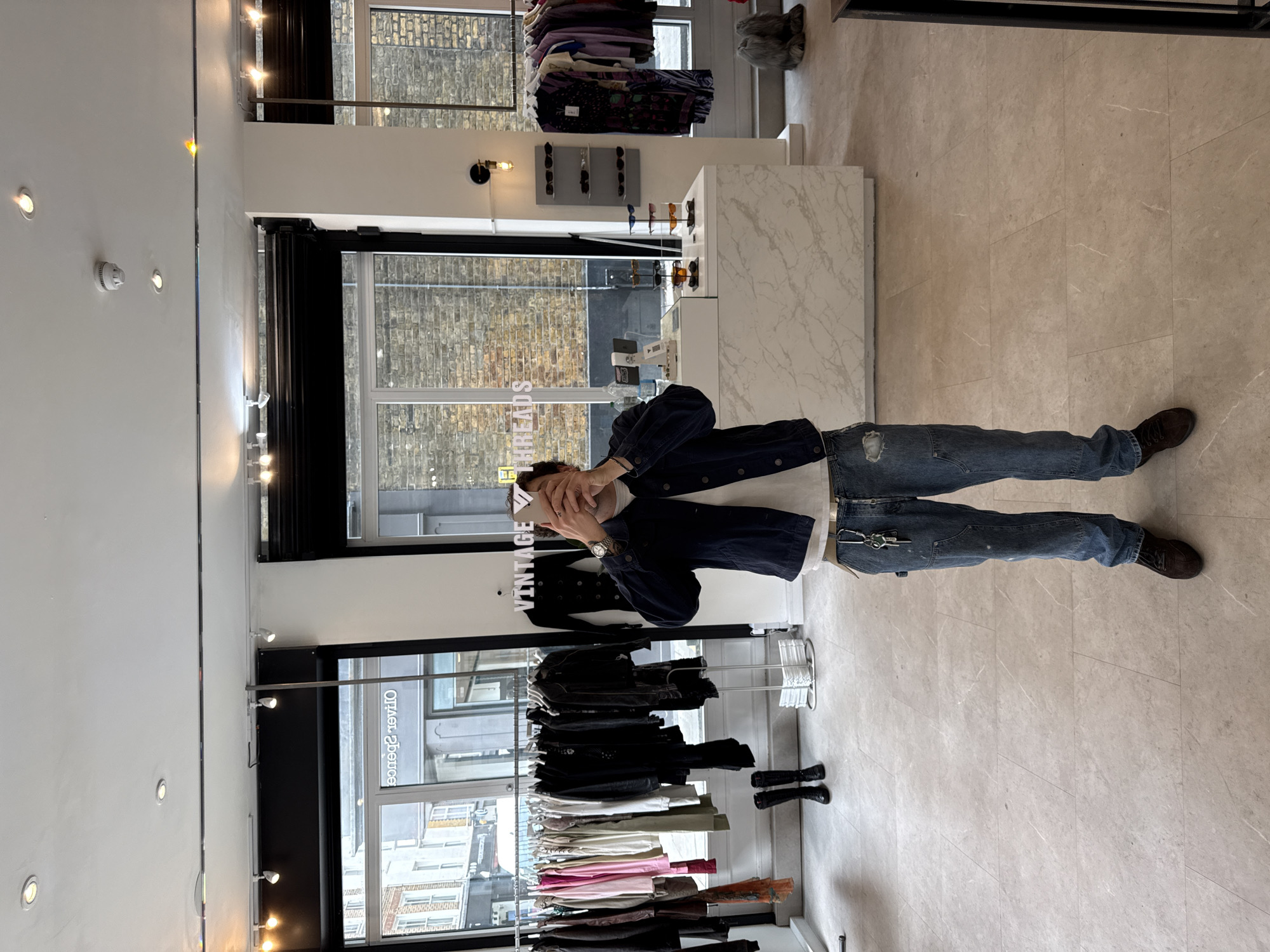 The image captures the interior of a brightly lit retail store, likely a fashion boutique, during the daytime. A male figure stands centrally, his face partially obscured by his hands or a phone he holds. He is dressed in a dark denim jacket, a light-colored t-shirt, blue jeans with a ripped knee on one leg and a carpenter's loop with keys clipped to it on the other, and dark shoes.
The store's interior features white walls, a light-colored tiled floor with faint white veining, and large windows that span a significant portion of one wall, allowing natural light to enter. Through these windows, an urban landscape with a brick building and glimpses of a street are visible.
Merchandise within the store includes clothing racks displaying a variety of garments, from dark jackets and tops to lighter colored and more colorful items. White, marble-patterned display counters hold rows of sunglasses. Recessed ceiling lights and additional track lighting illuminate the space and displays. A smoke detector is also visible on the ceiling.
Visible text includes "VINTAGE THREADS" reflected in reverse on an interior display, and "VINTAGE" clearly written on a white hanger or sign near the person. Through the window, the reversed text "OLIVER SPENCE" can be seen, likely a neighboring business sign. The setting is clean and modern, consistent with a contemporary shopping experience in an urban environment like London.