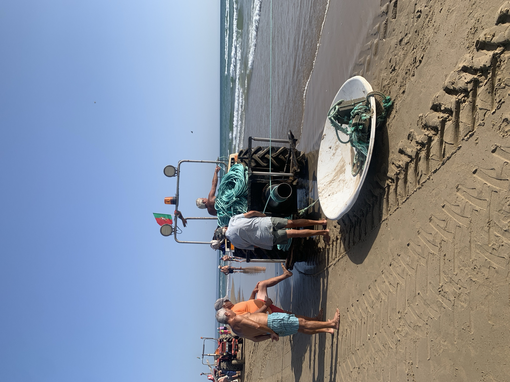 In Costa da Caparica, Portugal, on a sandy beach adjacent to the ocean with moderate waves under a clear blue sky, a group of men are gathered around a tractor. The tractor, which appears to be in operational condition, is positioned near the water's edge. It has large tires with distinct tread patterns imprinted on the wet sand. A significant amount of thick, coiled green rope is loaded onto the back of the tractor. A Portuguese flag is attached to the tractor's frame. In the foreground, a white, boat-like object, possibly a dinghy or a trough, rests on the sand, also with green rope secured to it. Several men are dressed in swimwear and casual attire, some are standing and observing, while others appear to be engaged in tasks related to the tractor and the rope. Another tractor is visible further down the beach. The scene suggests preparation or activity related to marine operations or beach maintenance.