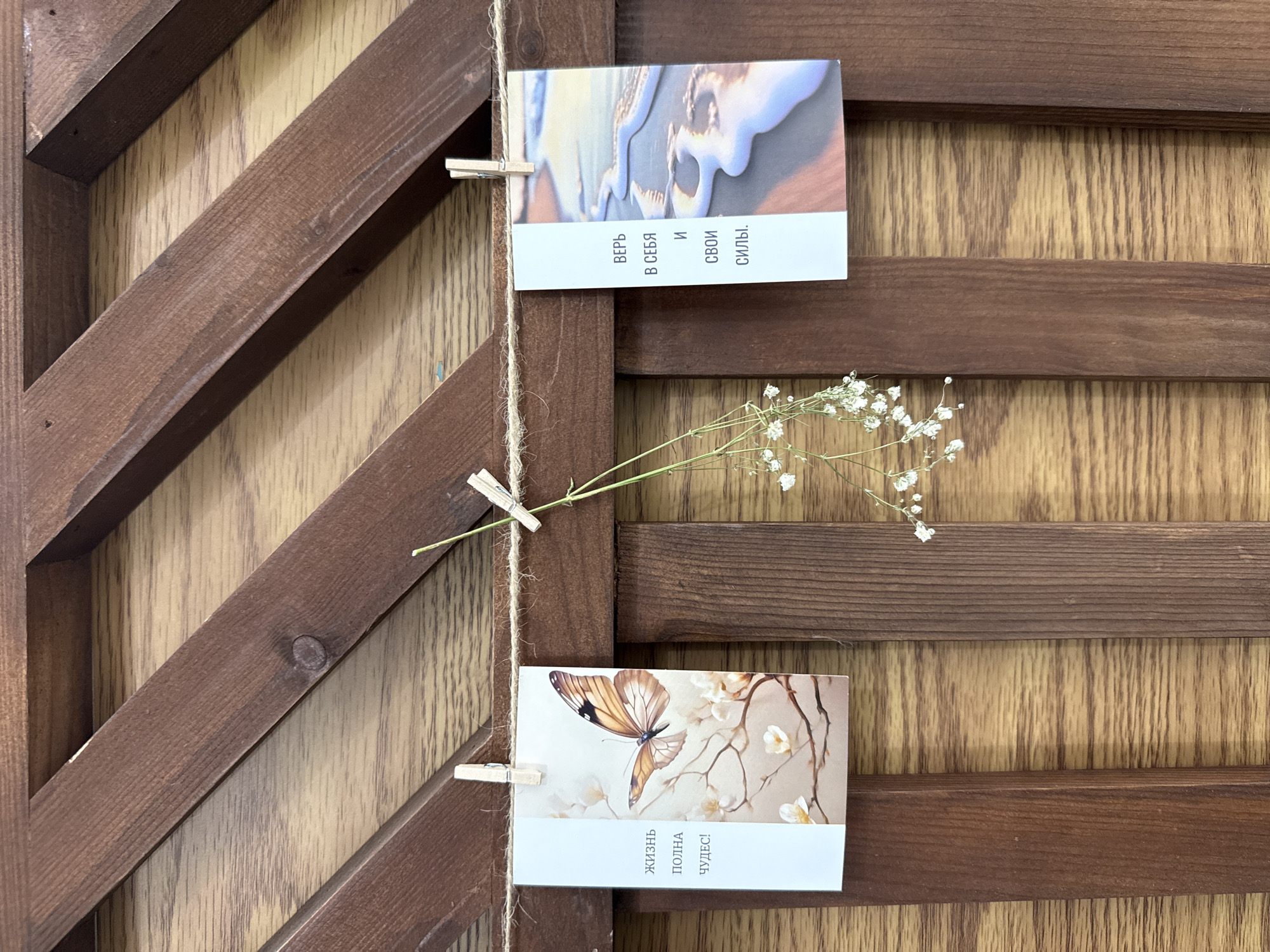 The image shows a close-up of a wooden lattice fence in Votkinsk, Russia. Two postcards are clipped to a piece of twine strung across the fence. The postcard on the left features an image of a butterfly on a branch with blossoms and the text "Жизнь полна чудес!" which translates to "Life is full of wonders!". The postcard on the right has an image of a beach at sunset and the text "Верь в себя и свои силы." which translates to "Believe in yourself and your strength.". A sprig of small white flowers hangs from the twine between the two postcards. The wooden fence has a warm, reddish-brown tone and a visible wood grain. The lighting suggests it is daytime.