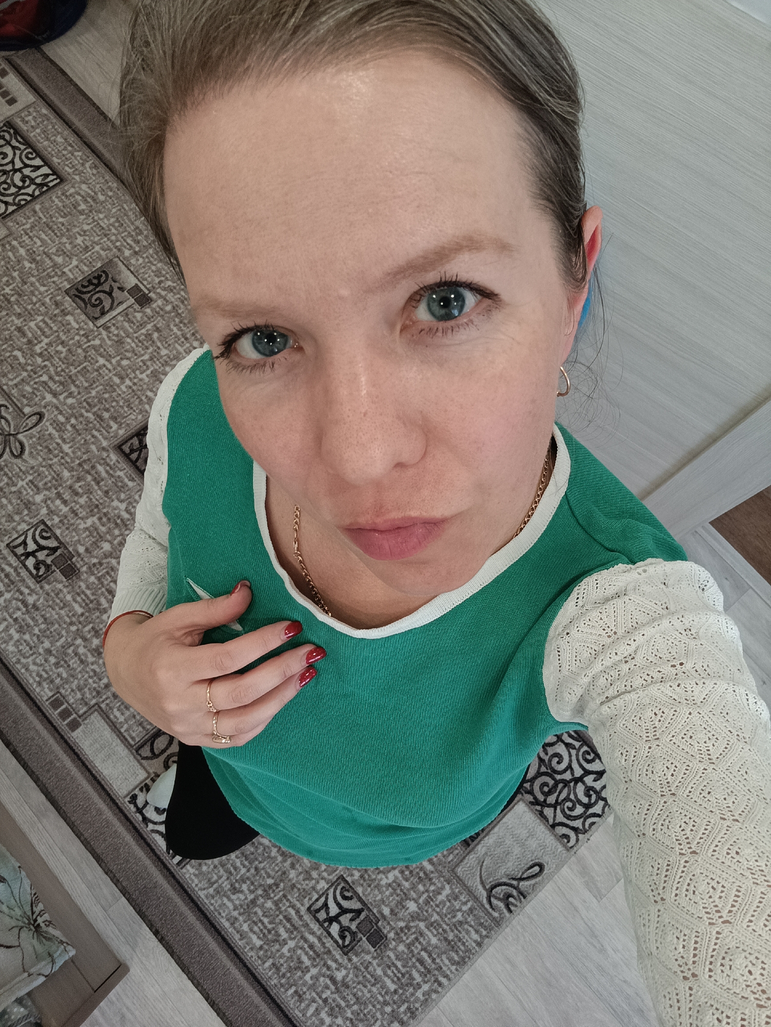 A woman with blue eyes and fair skin, wearing a green top with white lace sleeves and a gold necklace, looks up at the camera. She has short, greyish hair and her nails are painted red. She is wearing gold rings on her left hand, which she is holding to her chest. The background includes a patterned carpet and a light-colored wooden floor. The location is Chaikovsky, Russia.