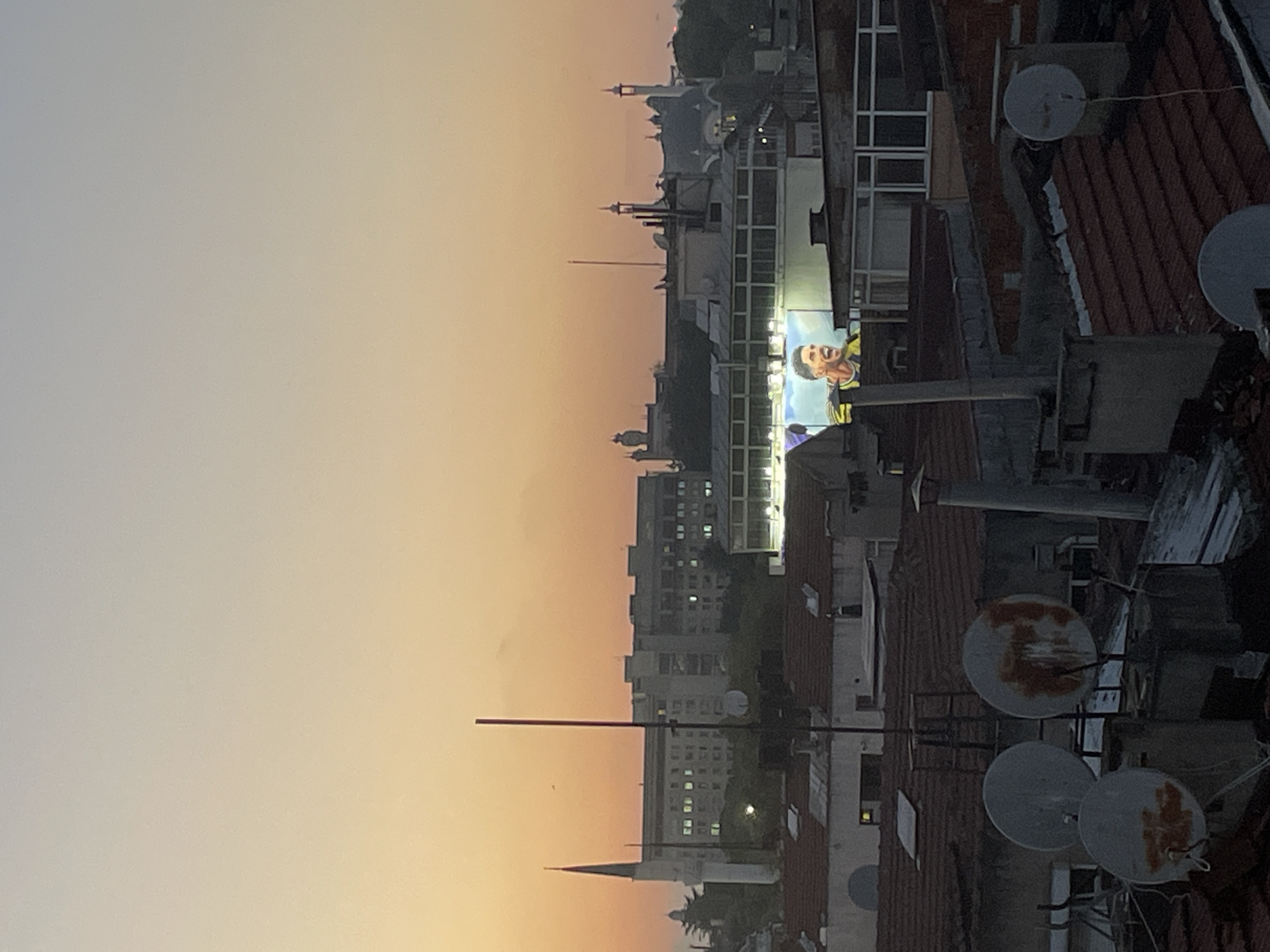 This is a rooftop view of Kadıköy, Turkey, at dusk. The sky is a gradient of orange and blue, with the sun setting on the horizon. In the foreground, several satellite dishes are visible on rooftops, some showing signs of rust.
Further in the distance, a large billboard featuring a close-up of a man in a yellow and black sports jersey is illuminated. The man has short dark hair and is shouting with his mouth open, his eyes wide. Behind the billboard, several buildings of varying architectural styles are visible, including a building with a tall, slender minaret to the left. Other structures in the background appear to be residential buildings and possibly commercial establishments. The overall scene depicts a bustling urban environment under a dramatic twilight sky.