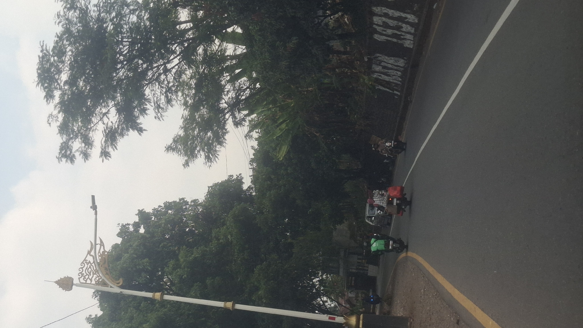 In the city of Cilaku, Indonesia, a street scene unfolds under an overcast sky. A lamppost with ornate, golden decorative elements stands prominently on the left side of the road, its white pole contrasting with the lush green foliage of trees lining the street. The road itself is asphalt, marked with a yellow line on the left and a white line in the center, indicating traffic lanes.
Several motorbikes are present. One, in the foreground, is ridden by a person wearing a green jacket, with a delivery box mounted on the back. Further down the road, another motorbike carries two people, one of whom appears to be wearing a headscarf and is also transporting items in red bags. A third motorbike is visible further still, with a single rider. A car is also present on the road, and a dark-colored vehicle can be seen on the left side of the road.
The background is dominated by dense green trees, some with large leaves resembling banana leaves. A low wall runs along the right side of the road, covered with what appears to be graffiti or markings, including numbers that resemble "1933". The overall impression is a typical street scene in a tropical environment, with a mix of vehicles and natural elements.