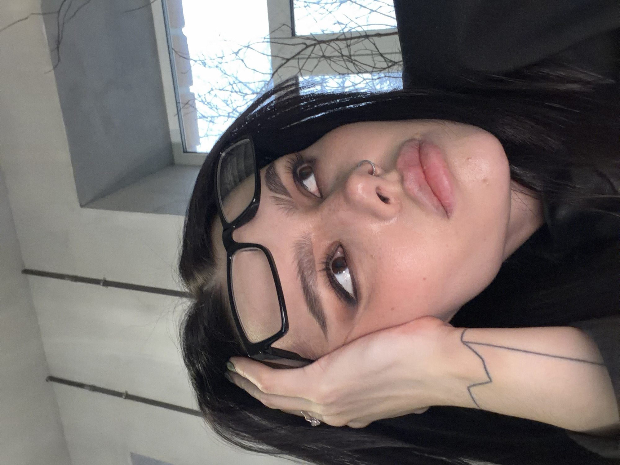 A woman with dark hair and glasses is lying on her side, resting her head on her hand. She has a nose ring and a tattoo on her wrist. The background includes a window with branches visible outside. The location is not identifiable as Moscow, Russia, based on the image.