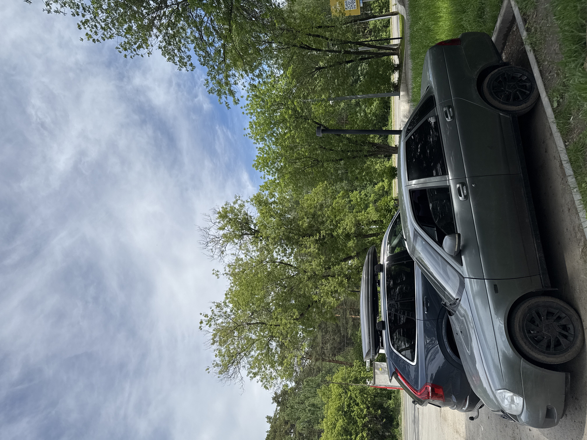 This outdoor daytime image, oriented vertically, depicts two cars parked alongside a curb next to a lush green treeline, all beneath a partly cloudy sky.
In the foreground, a dark grey car, resembling a sedan or hatchback (possibly a Lada Priora model), is parked with its front facing towards the left side of the frame. It features black multi-spoke alloy wheels and tinted windows, with its front bumper, headlight, and right side visible. Behind it, a darker blue or black station wagon/SUV is parked, also facing left. This second vehicle is equipped with a grey roof cargo box and its red rear lights are visible.
The cars are situated on a paved surface, bordered by a concrete curb separating it from a strip of green grass. Beyond the grass, a dense expanse of deciduous trees with vibrant green foliage dominates the middle and left portions of the image, suggesting a park or forested area adjacent to the parking spot. Several slender, dark poles, possibly streetlights or utility poles, can be seen interspersed among the trees.
The upper left section of the image is filled with a bright blue sky streaked with scattered, wispy white clouds. In the far right background, partially obscured by the trees, a yellow sign with a visible QR code and some illegible text below it can be discerned.
The scene conveys bright daylight and clear weather. There is no specific activity or event taking place, just stationary vehicles in what appears to be a parking area or roadside in Perm, Russia, adjacent to a green space.
