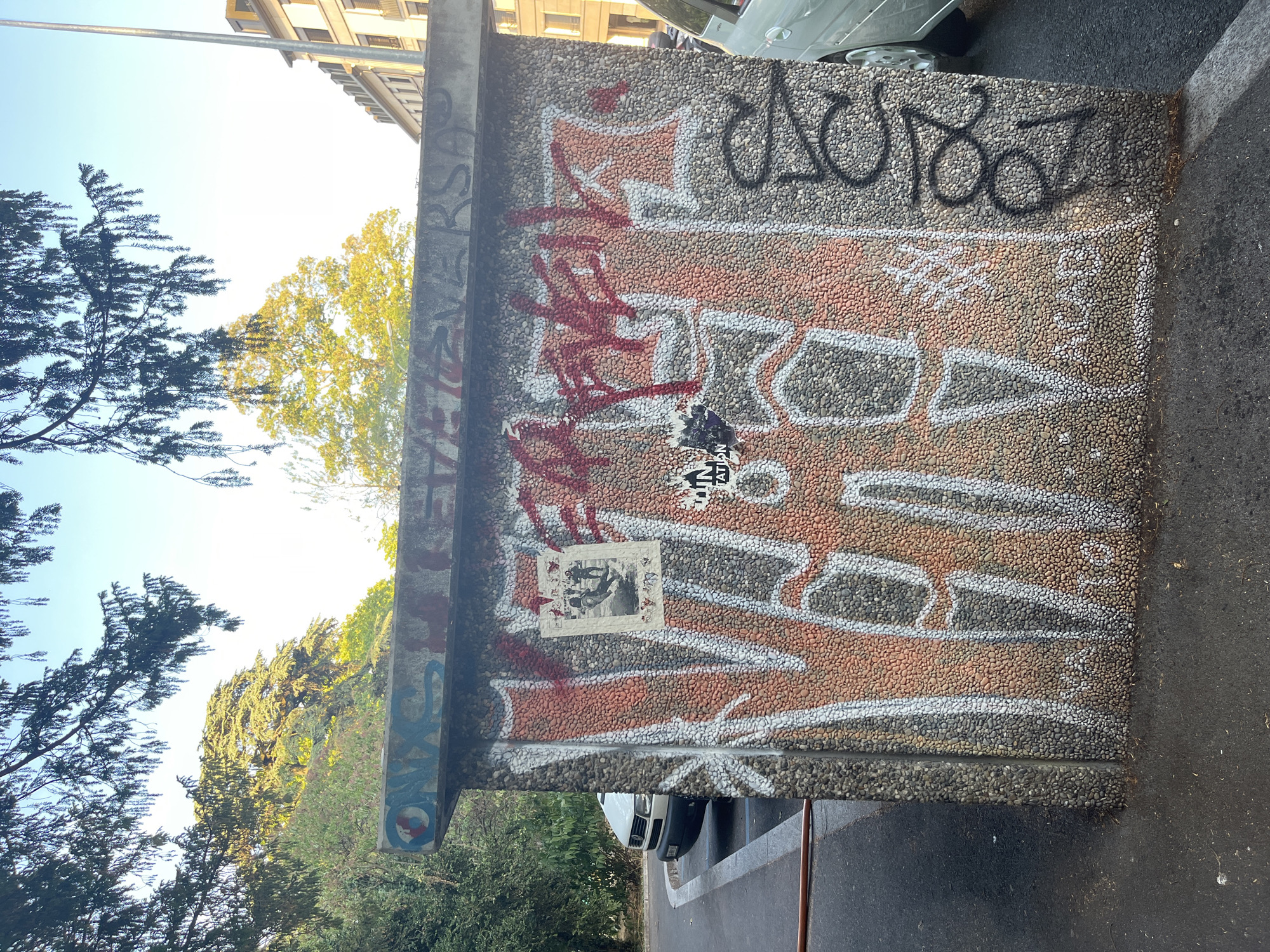 A concrete structure, possibly a utility box or a small building, is covered in graffiti. The structure's exterior is a mosaic of small, rough pebbles. Large, stylized graffiti in red and white paint covers most of the visible surfaces. A poster depicting two figures in a dynamic pose is affixed to the left side of the structure. To the right of the poster, a smaller black and white sticker is visible. Black graffiti in a circular, stylized script is present on the right side of the structure. The structure is situated on an asphalt surface, likely a sidewalk or road. To the left, a white van is parked. In the background, trees with green and yellow foliage are visible against a clear blue sky. Further in the background, a multi-story building with light-colored facades and visible windows can be seen. The image was taken outdoors, likely in an urban environment in Geneva, Switzerland, given the context.