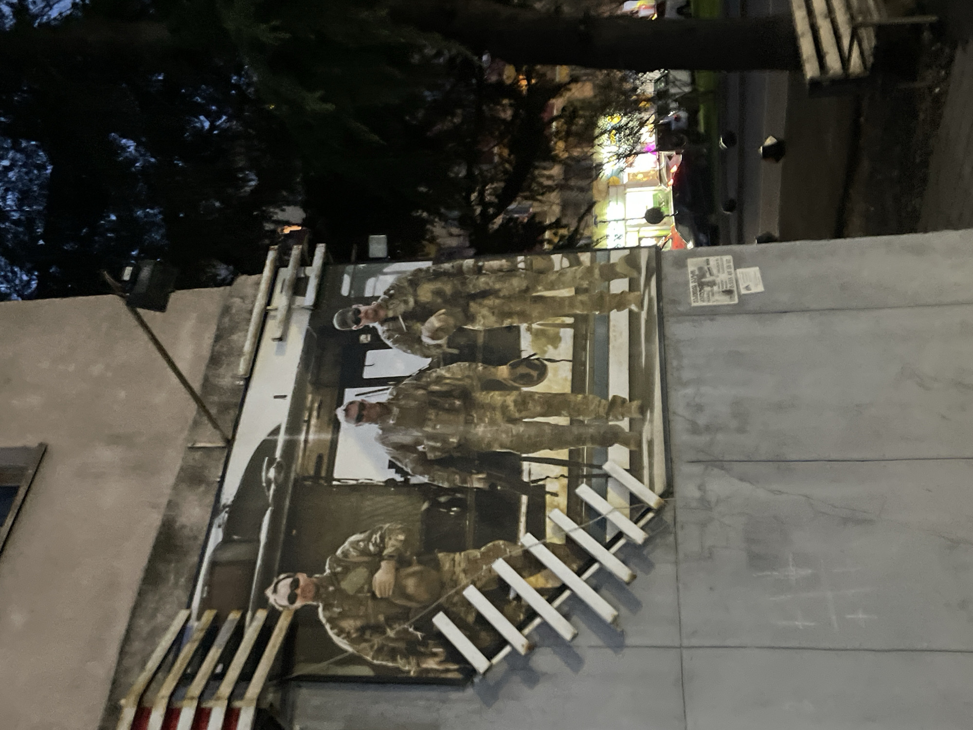 The image displays an outdoor scene at dusk or night in Tbilisi, Georgia, featuring a large vertical poster mounted on a light-colored wall. The poster depicts three individuals in full military camouflage uniforms, helmets, and sunglasses. They appear to be soldiers, standing within what looks like the interior of a military vehicle or structure, characterized by visible metal framework.
In front of the poster, a barrier or fence composed of white horizontal slats is visible. Beneath these slats, a pattern of red and white stripes suggests a flag, possibly the Georgian flag. To the right of the poster, on an adjacent wall section, faint chalk drawings resembling tic-tac-toe grids are present.
Above the poster, the silhouettes of dark tree foliage are visible against the dark sky. Further in the background, soft, colorful lights indicate a lively area such as a street with shops or entertainment venues. Several small flyers or papers are taped to the wall above the poster; partial text on one includes "GLOBAL..." and numbers like "47 85 92" and "100000," too blurry for full legibility. The scene captures a public display with military imagery in an urban environment.