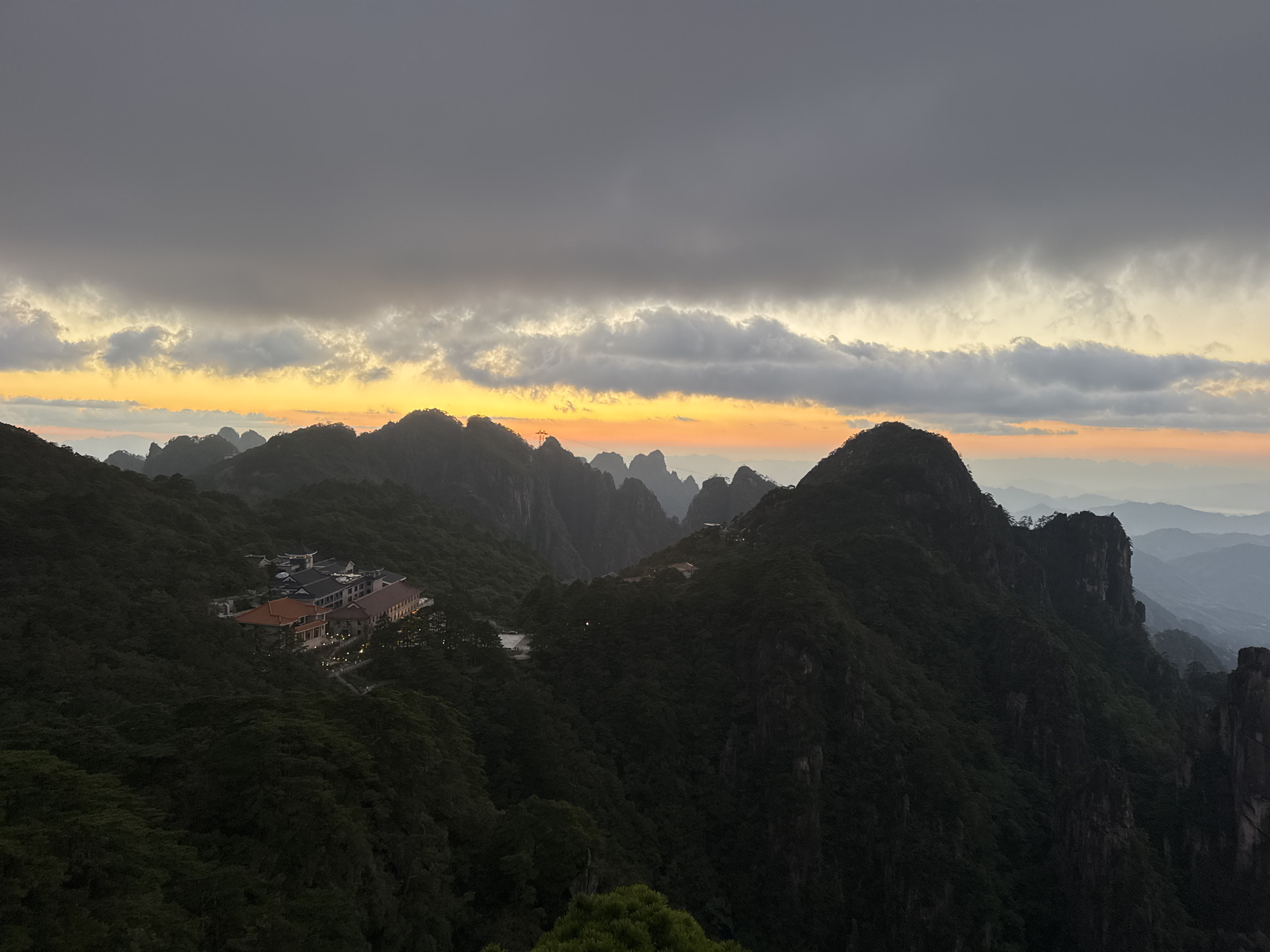This is a high-angle, wide-shot photograph capturing a mountain landscape at dusk, likely in Huangshan District, China. The sky is dominated by dark, brooding clouds in the upper half, transitioning to a bright band of orange and yellow light near the horizon, indicating the setting or rising sun. Below the sky, a series of steep, tree-covered mountain peaks recede into the distance. In the mid-ground, nestled on a mountainside, is a complex of buildings with red-tiled roofs, appearing to be a resort or hotel. The surrounding terrain is densely forested with dark green foliage. The overall scene evokes a sense of dramatic natural beauty, heightened by the interplay of light and shadow. There are no people or animals visible in the image.