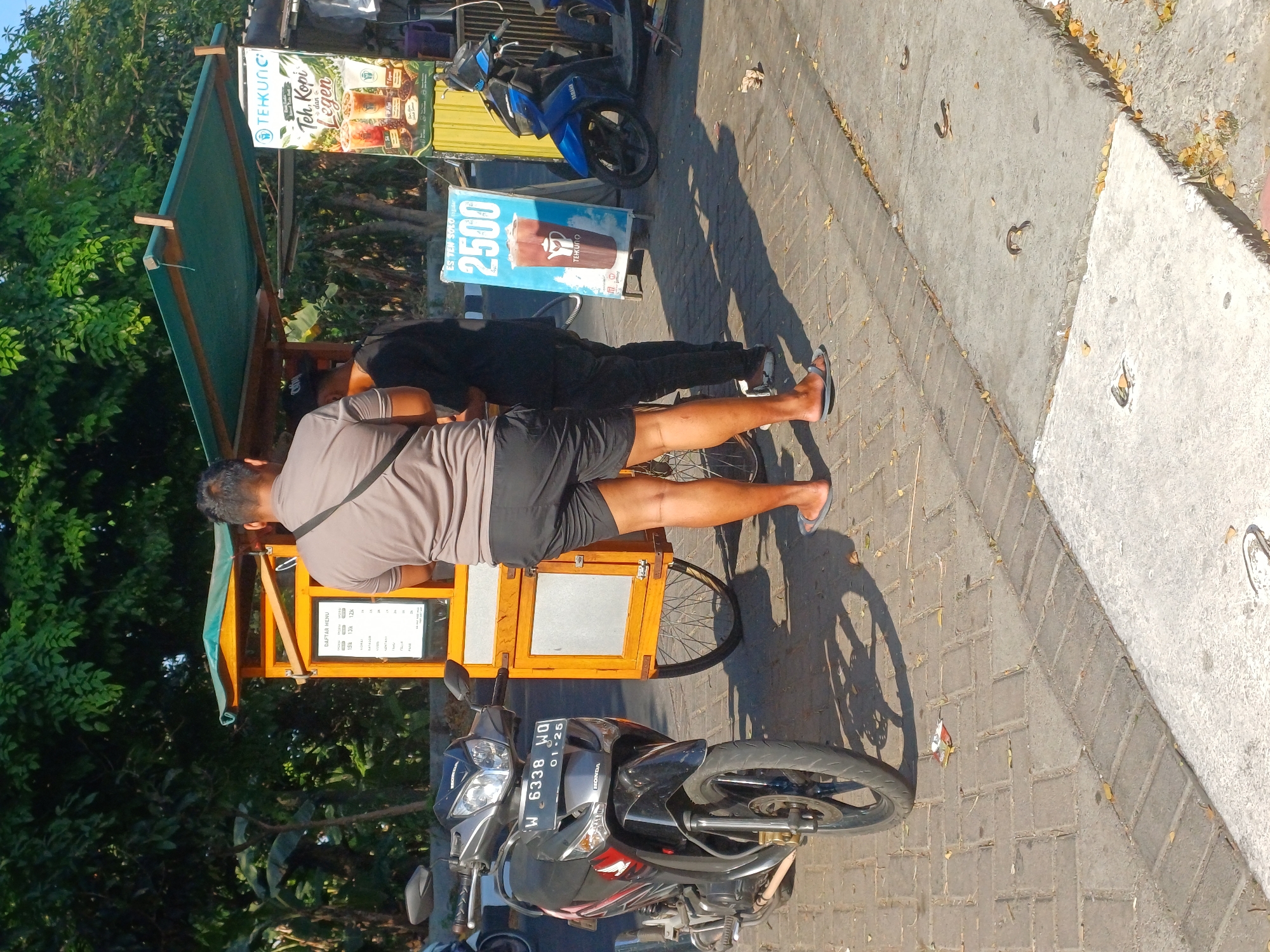 The image depicts a street scene in Pulungan, Indonesia, on a sunny day. A mobile food cart, predominantly yellow with a green awning, is set up on a paved sidewalk. A menu is displayed on the side of the cart. Two men are standing near the cart. One man, wearing a gray t-shirt and black shorts, stands with his back to the camera, facing the cart. The other man, wearing a black shirt and dark pants, is partially visible behind him. A motorcycle, black and red with a license plate reading "W 6338 WO 01-25", is parked to the left of the cart. A blue motorcycle is parked to the right, behind a bright blue advertising sign that displays a drink and the text "ES TEN SOLO 2500 TEKUNO". Trees and foliage form a backdrop to the scene. The ground is paved with interlocking bricks and concrete sections, with scattered fallen leaves. The lighting suggests it is midday, casting distinct shadows.