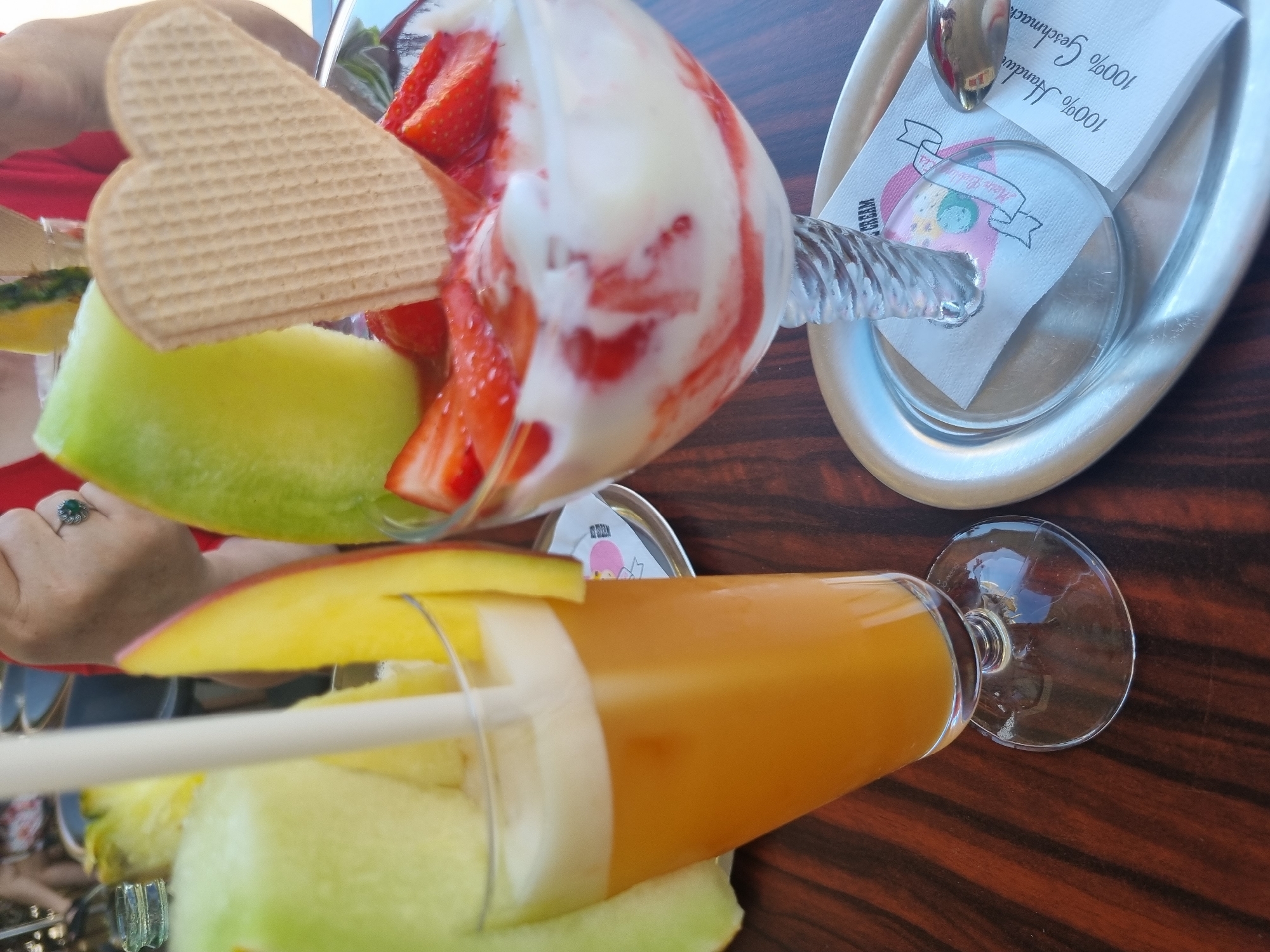 The image depicts a table at a cafe, possibly in Saarbrücken, Germany. There is a dessert glass with strawberries, ice cream and a heart-shaped wafer cookie. The glass is partially obscured by a slice of melon. Beside it, there's a glass of orange juice with melon slices and mango garnish. A silver tray is visible, holding napkins, a logo of "Heiss Heiss Eis", and a spoon. The table is wooden and has a dark wood grain. A woman's hand with a ring is visible near the dessert glass. The scene suggests a relaxing moment at a cafe.