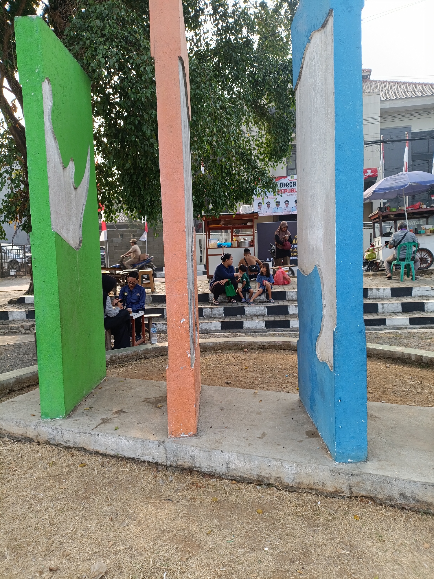 This image depicts an outdoor scene, likely in the city of Cariu, Indonesia, featuring a public space with several individuals. In the foreground, three large, colorful pillars stand prominently: one green, one orange, and one blue, with white sections on each. Behind the pillars, a group of people are seated on a set of stairs with black and white risers.
On the left side of the stairs, two adults and two children are gathered. One adult, wearing a dark headscarf and blue top, is seated, while another adult and two children are sitting in front of them. A red bag rests beside the group. To the right, another group of people are seated on the stairs. One person is sitting on a low stool, looking at their phone, while a child sits next to them. Further to the right, a person is seated on a green chair, facing away from the camera, near a food stall with a blue umbrella.
In the background, a banner with Indonesian text and images of men in uniforms is visible. Trees with dense foliage are present behind the pillars. A motorcycle is seen in the middle ground, ridden by a person wearing a light-colored shirt. The ground is covered with dry grass and dirt. The overall impression is a lively public gathering space.