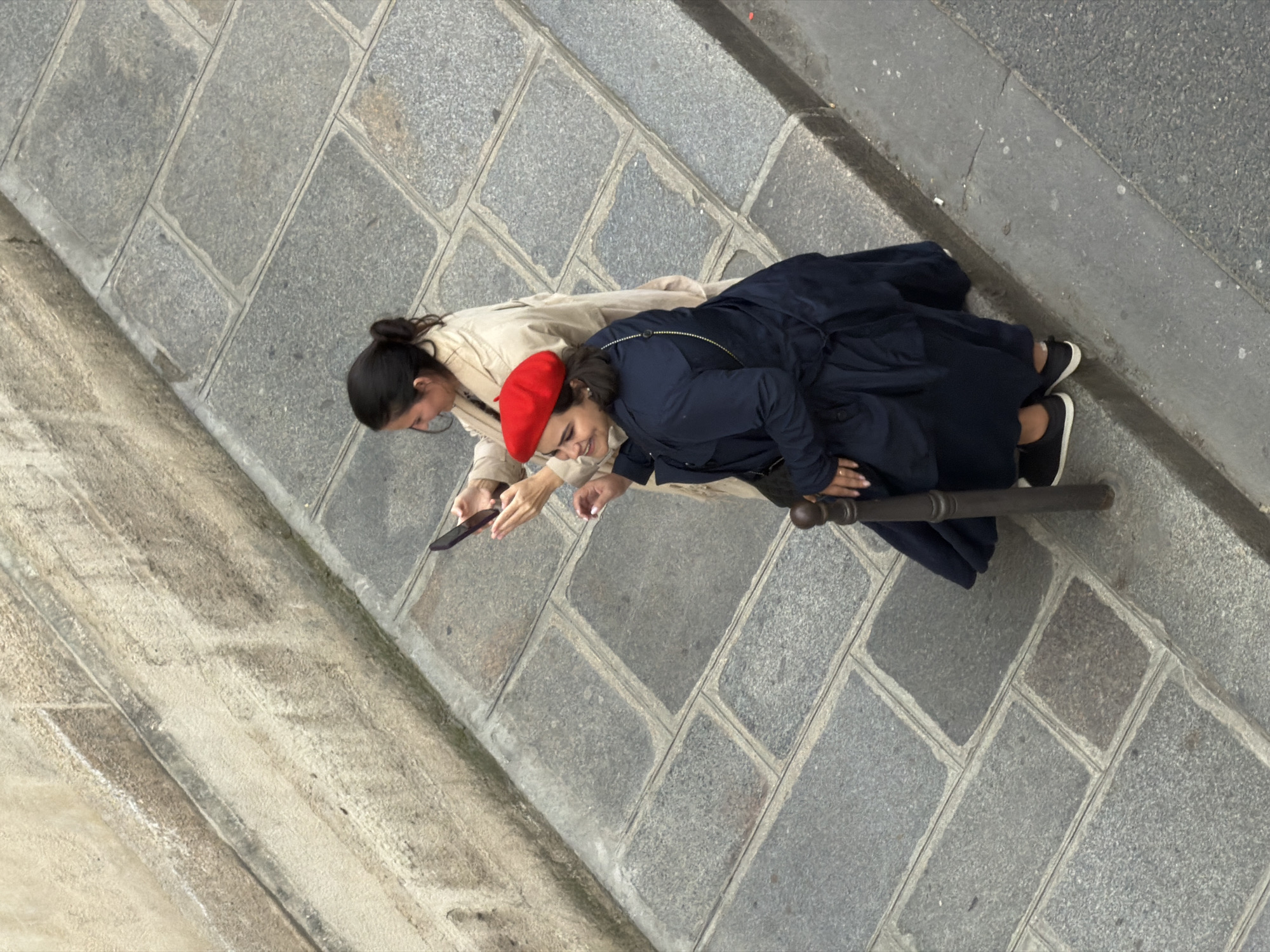 Two women are sitting close together on a gray cobblestone surface, possibly a sidewalk or terrace in Paris, France. One woman wears a red beret and a navy blue coat, while the other is dressed in a beige trench coat. The first woman is smiling. The other woman is looking at a phone. They are resting against a dark metal railing.